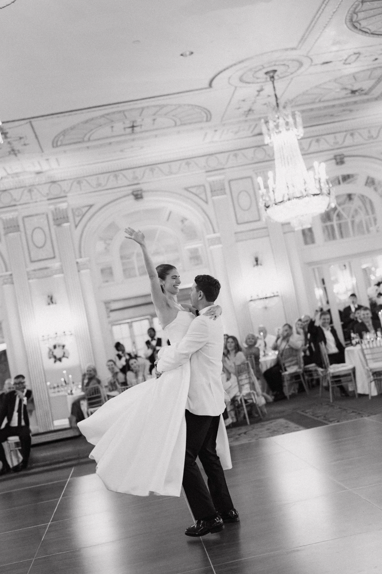 Black and white photo of a bride and groom dancing at a wedding reception. The bride is being lifted by the groom, smiling, with her arm raised. Guests are seated at decorated tables in a grand, ornate hall with chandeliers.
