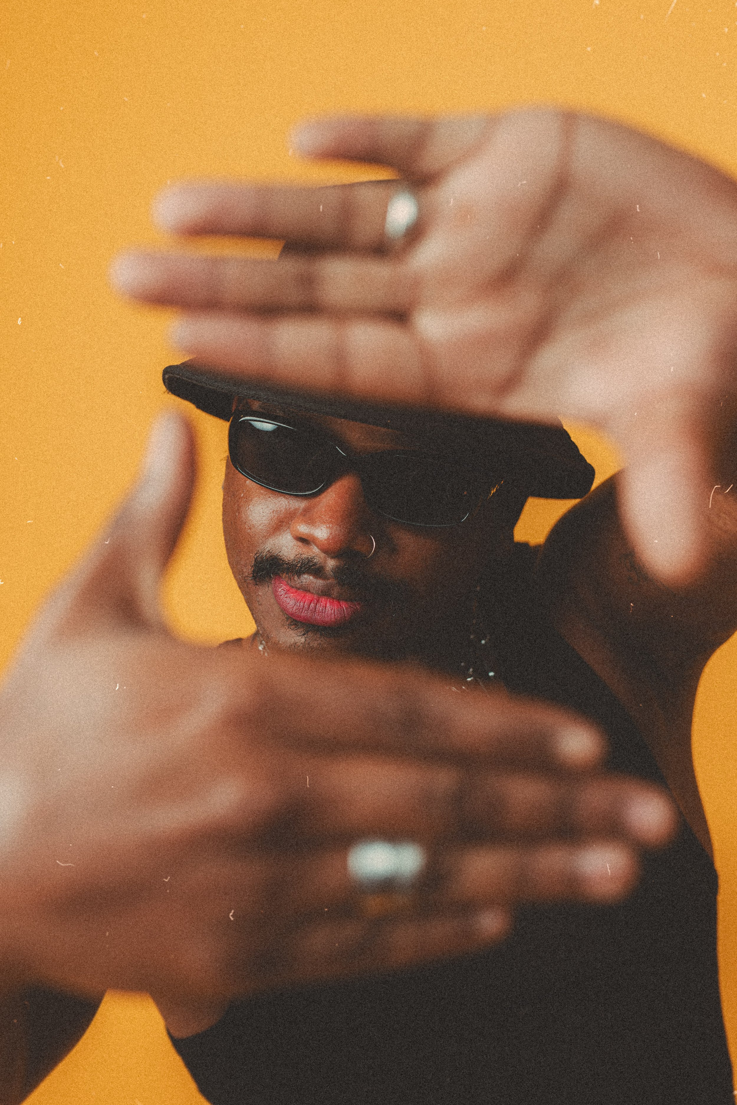 Person wearing sunglasses and a hat, framing face with hands, orange background.