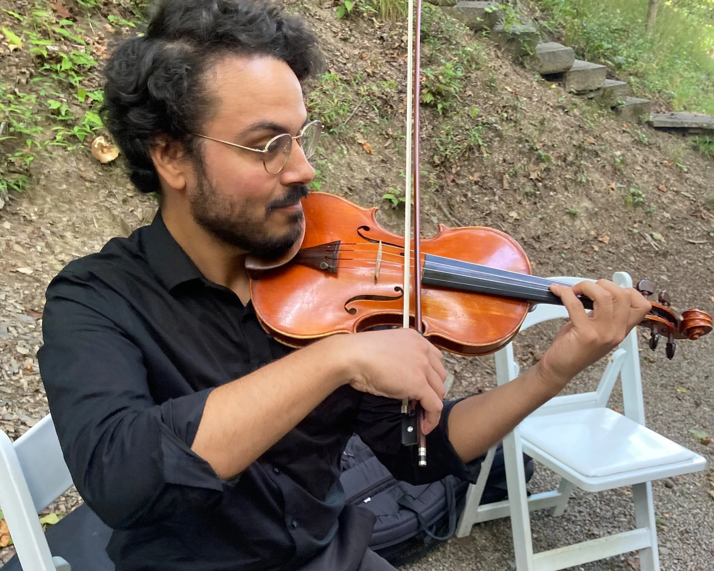 A man with curly dark hair, glasses, and a beard playing a violin outdoors, seated on a white folding chair with trees and steps in the background.