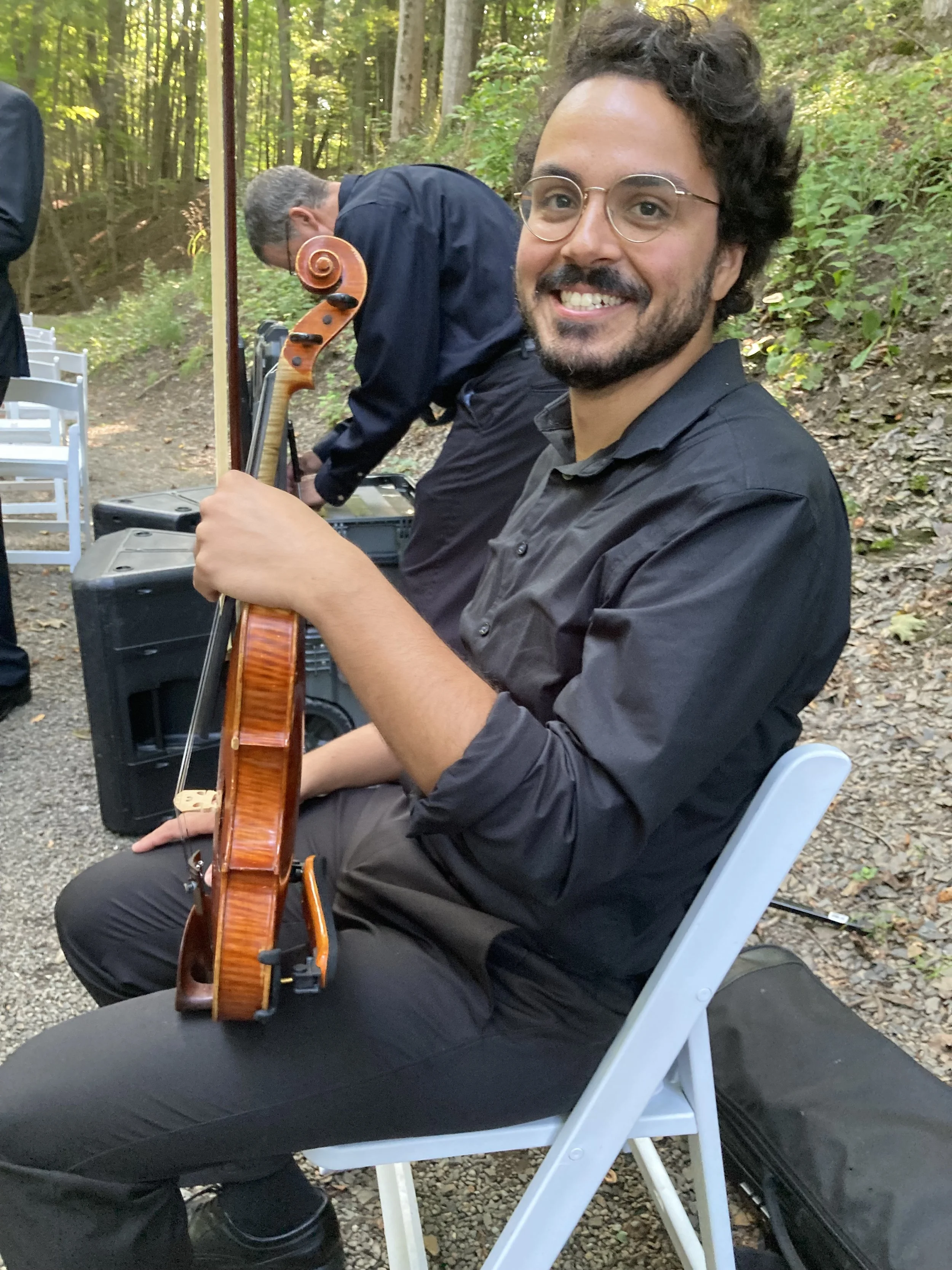 Reuben sits at an outdoor venue, holding his violin and smiling.