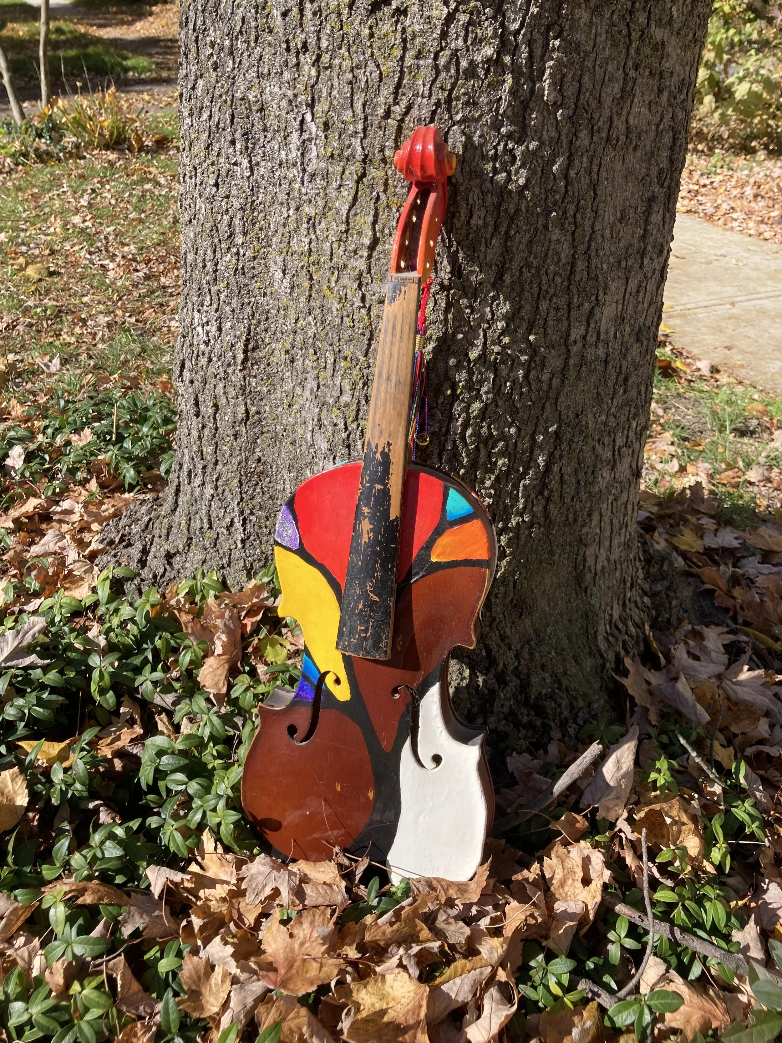 Colorful painted violin leaning against a tree trunk in an outdoor setting with leaves on the ground.