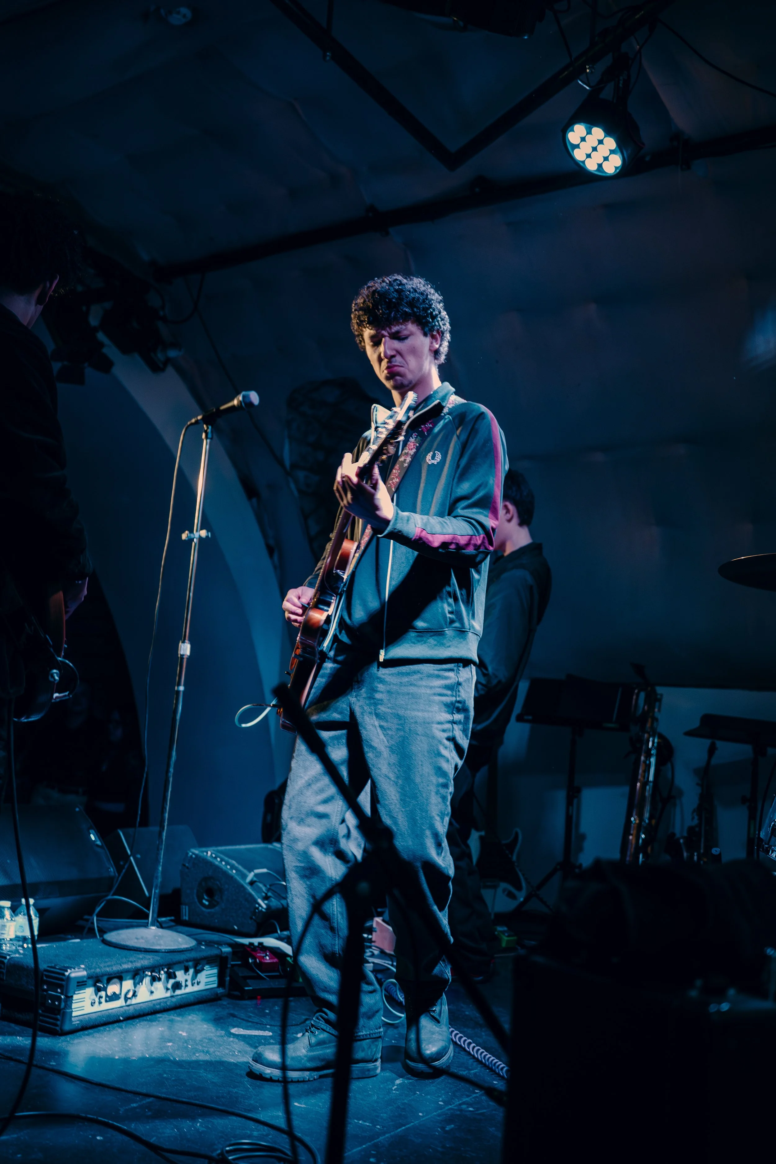 A young man with curly hair playing an electric guitar on stage during a live music performance, with stage lights illuminating him.