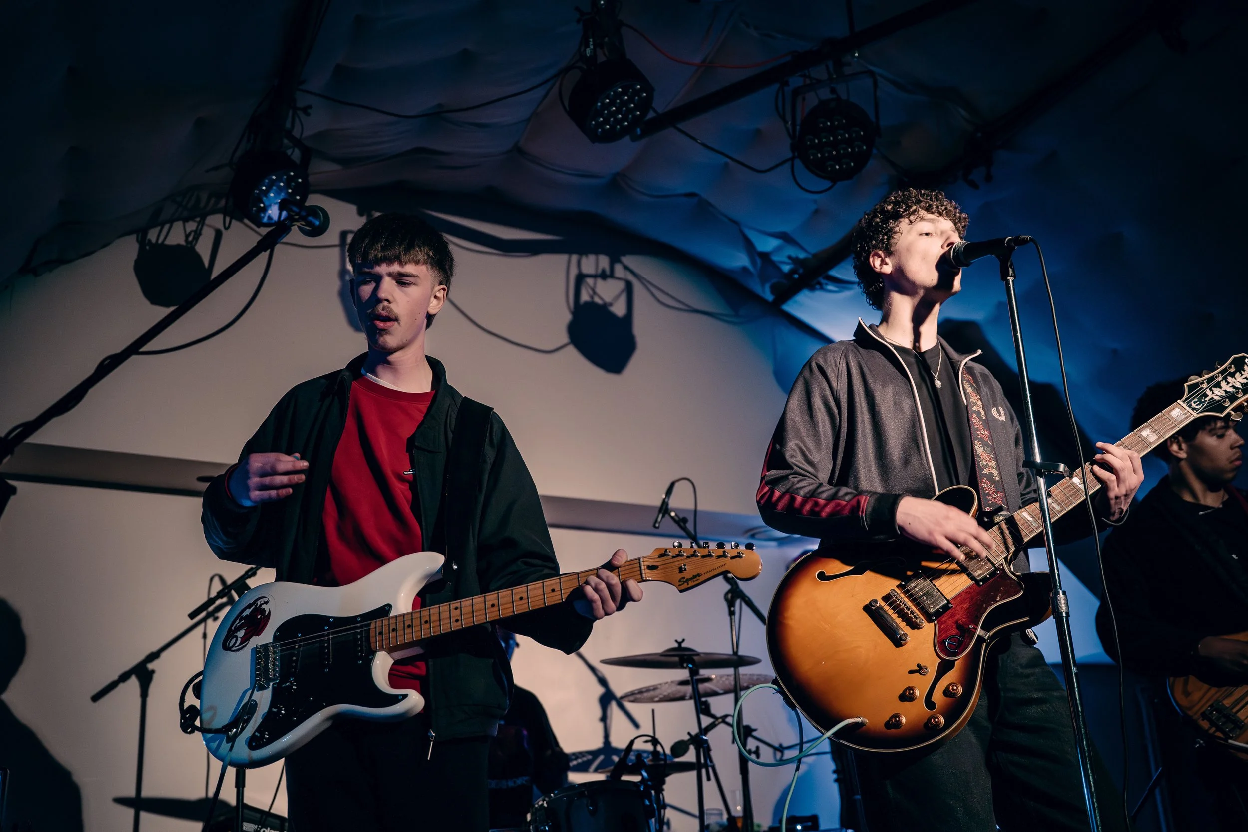 Two young men performing with electric guitars on stage, one singing into a microphone. Stage is lit with blue and white lights, with musical equipment in the background.
