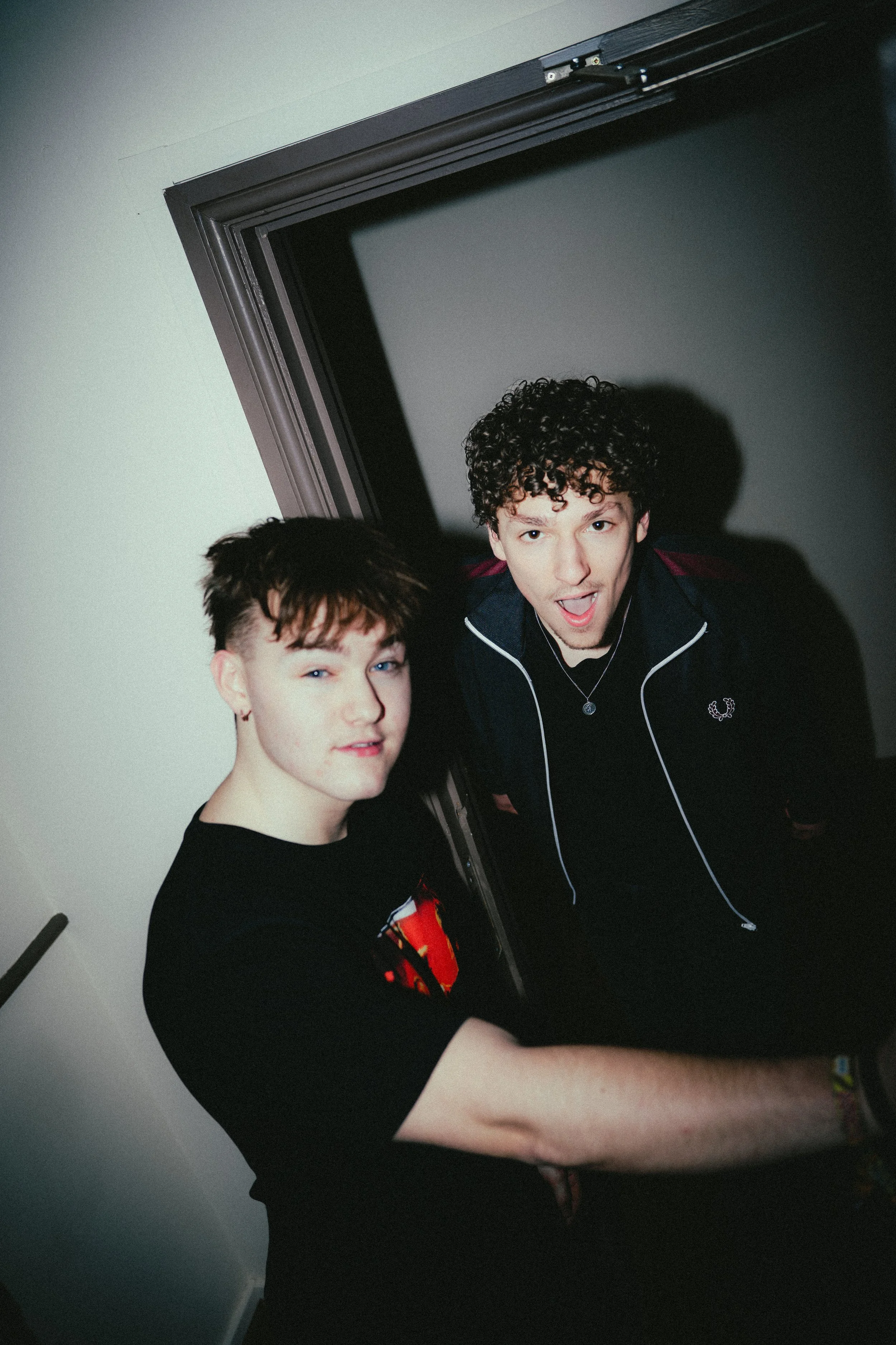 Two young men standing indoors near a doorway. One has short, dark hair and is winking slightly; the other has curly hair and is making a surprised or excited expression.