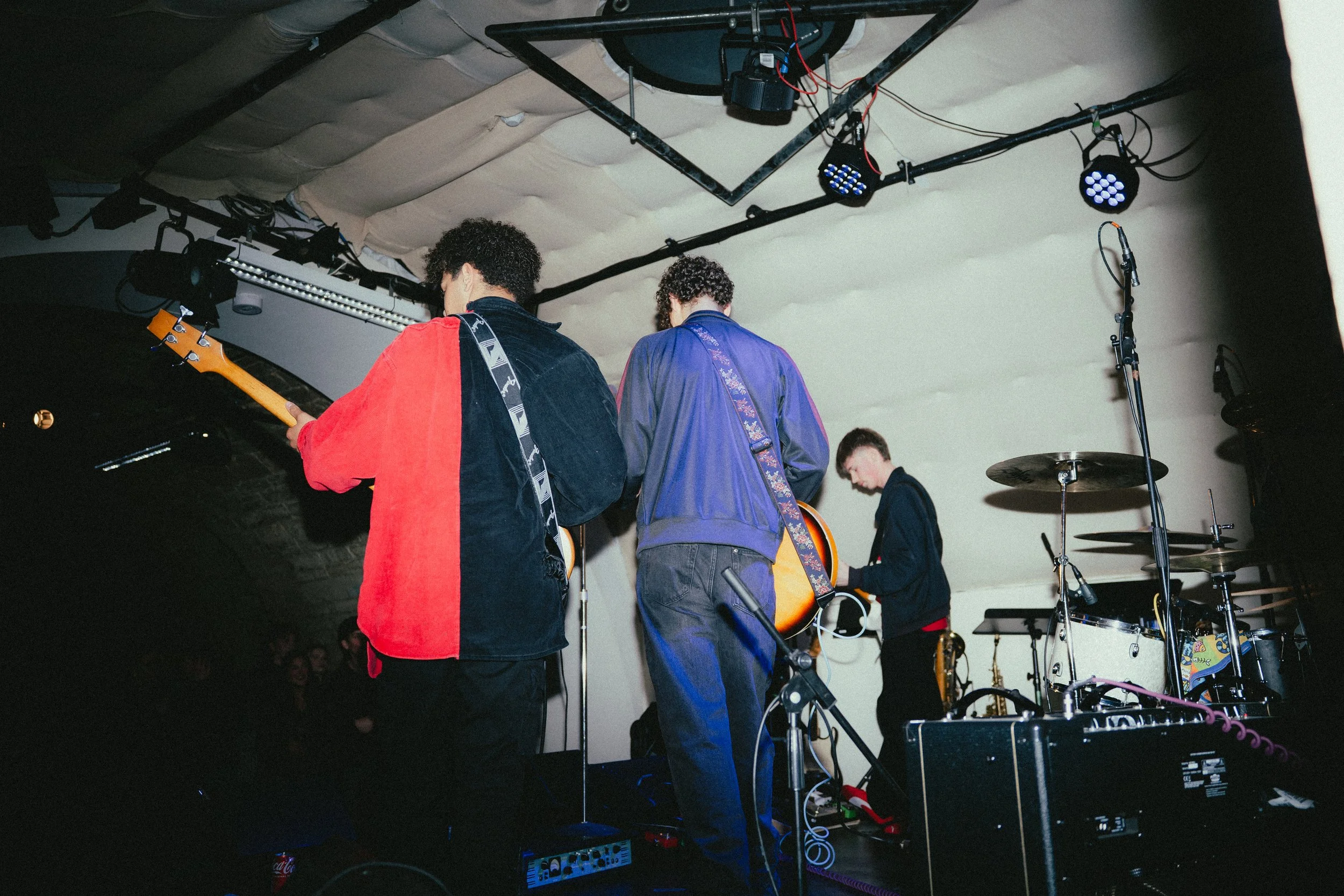 Three musicians on stage playing instruments, with a drum set visible on the right and stage lighting above.