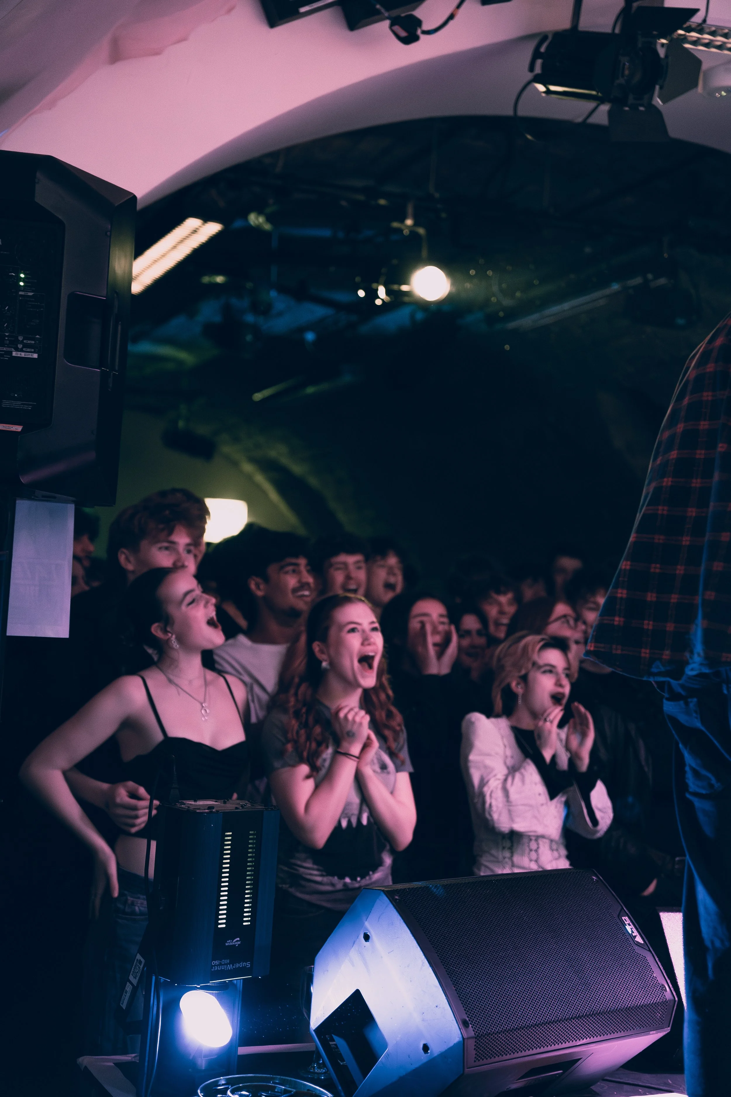 Audience at a concert or live show, reactions include laughter and excitement, stage equipment visible in foreground