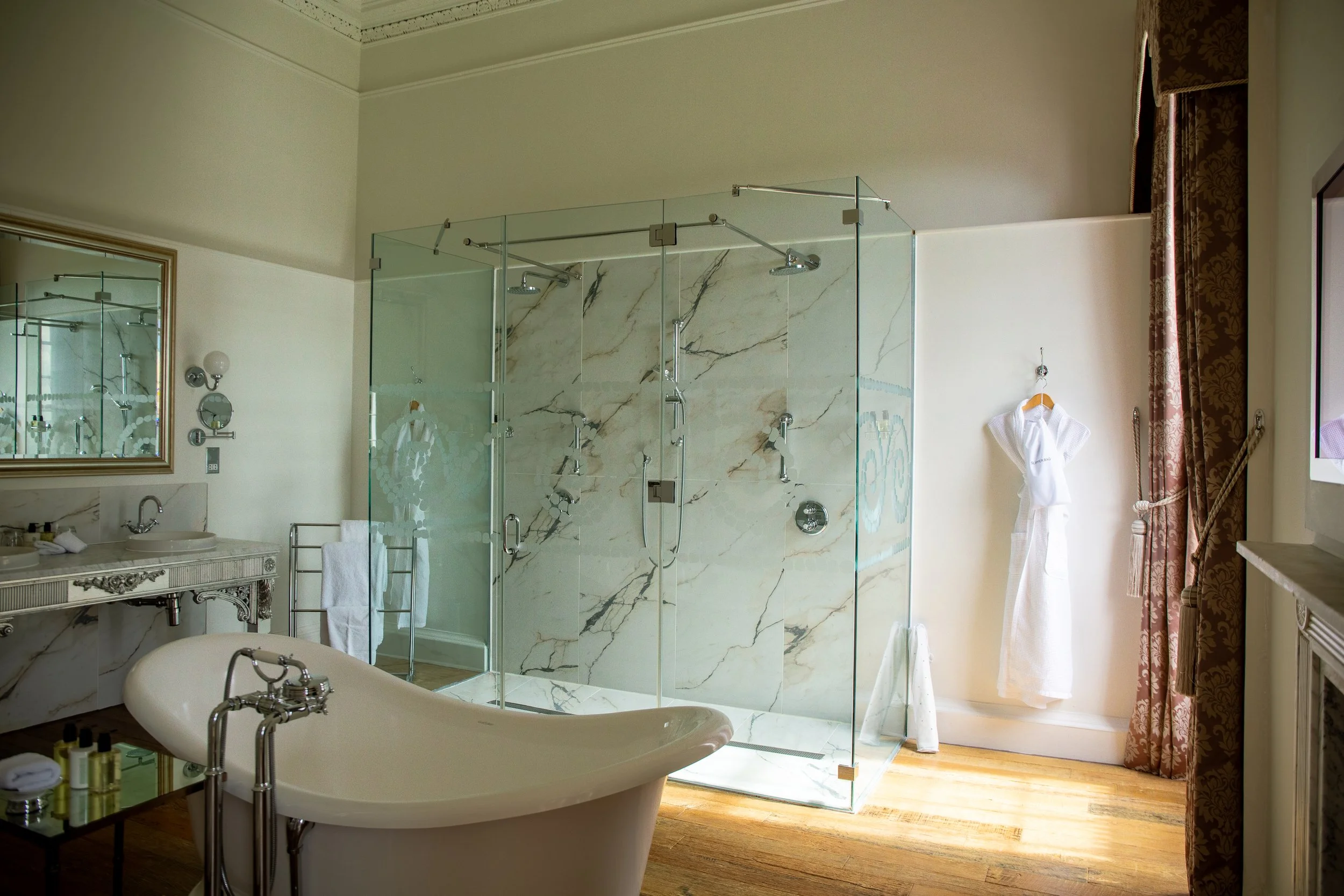 Luxurious bathroom with vintage white bathtub, glass-enclosed shower with marble walls, white robe hanging, and ornate mirror and furnishings.