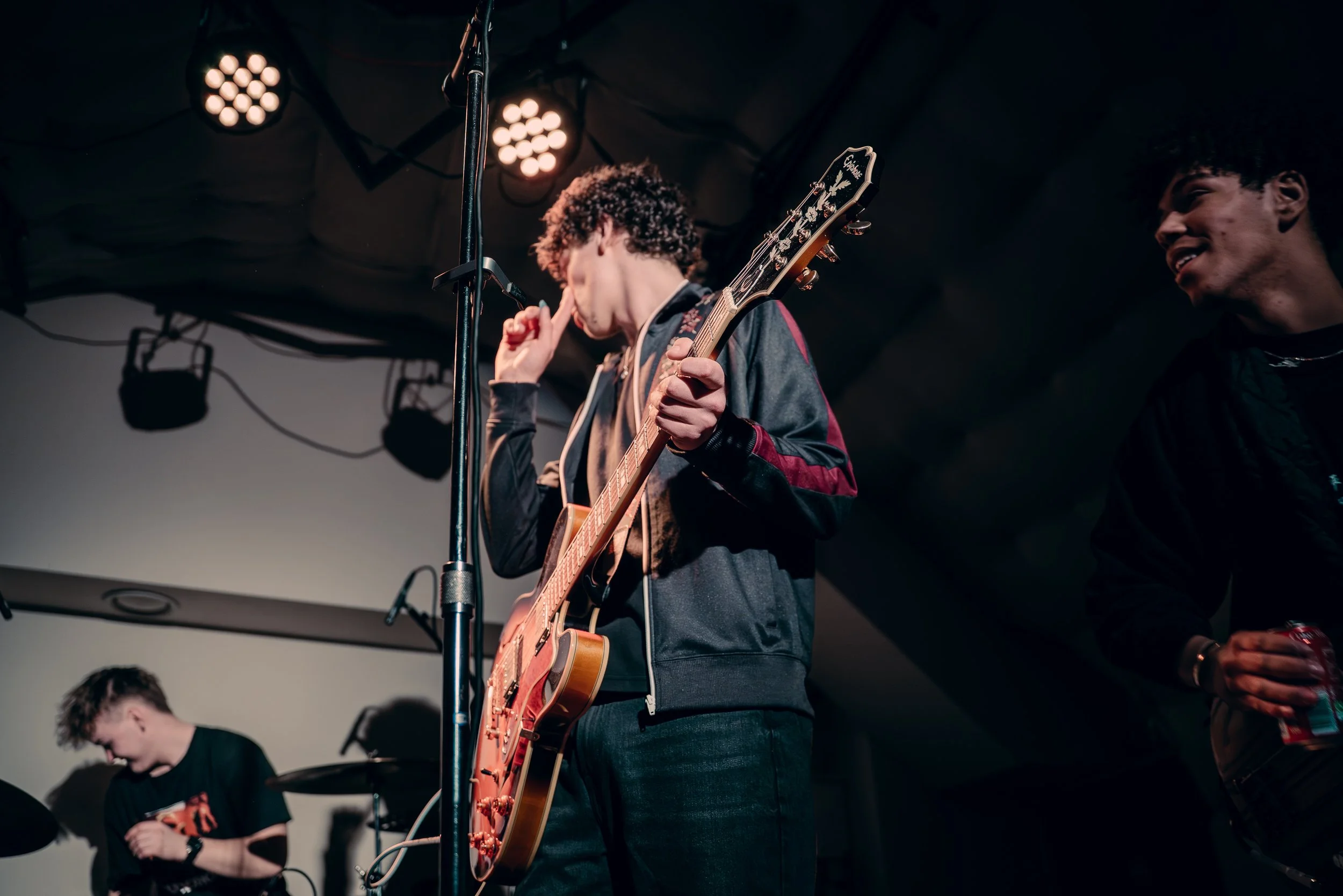 Three young men, one with a guitar, in a dimly lit room with stage lights.