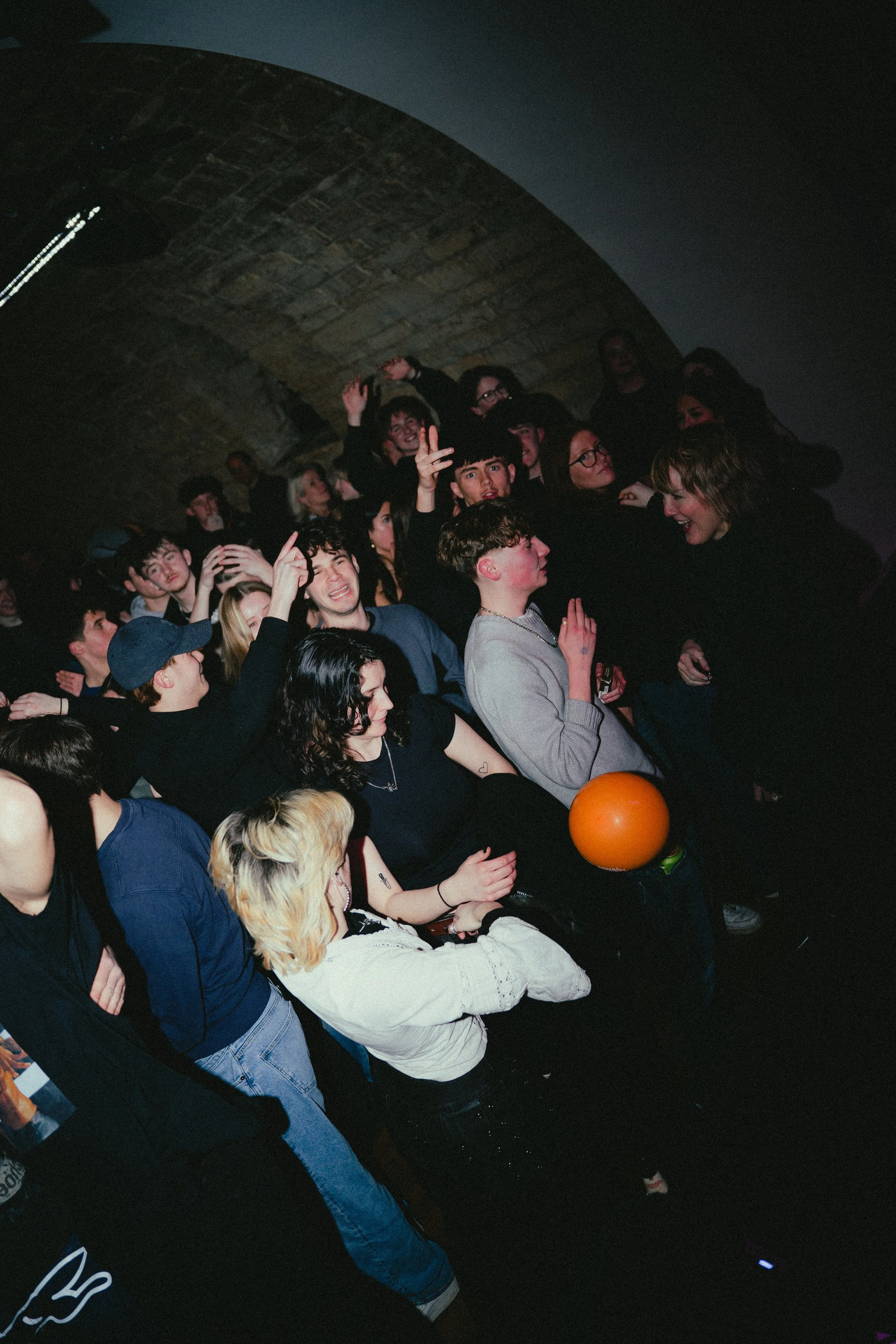 A crowd of young people at a concert or party, some dancing and smiling, with a brick arch ceiling in the background.