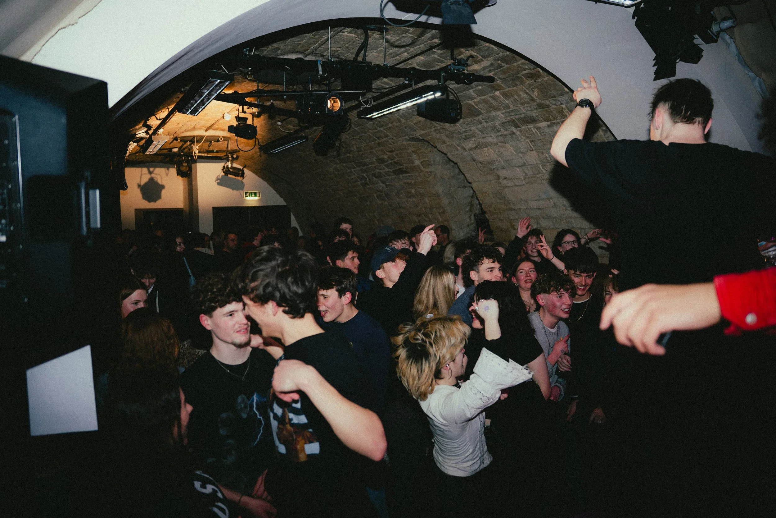 People enjoying a lively concert or party indoors under an arched stone ceiling, with some raising their hands and smiling.
