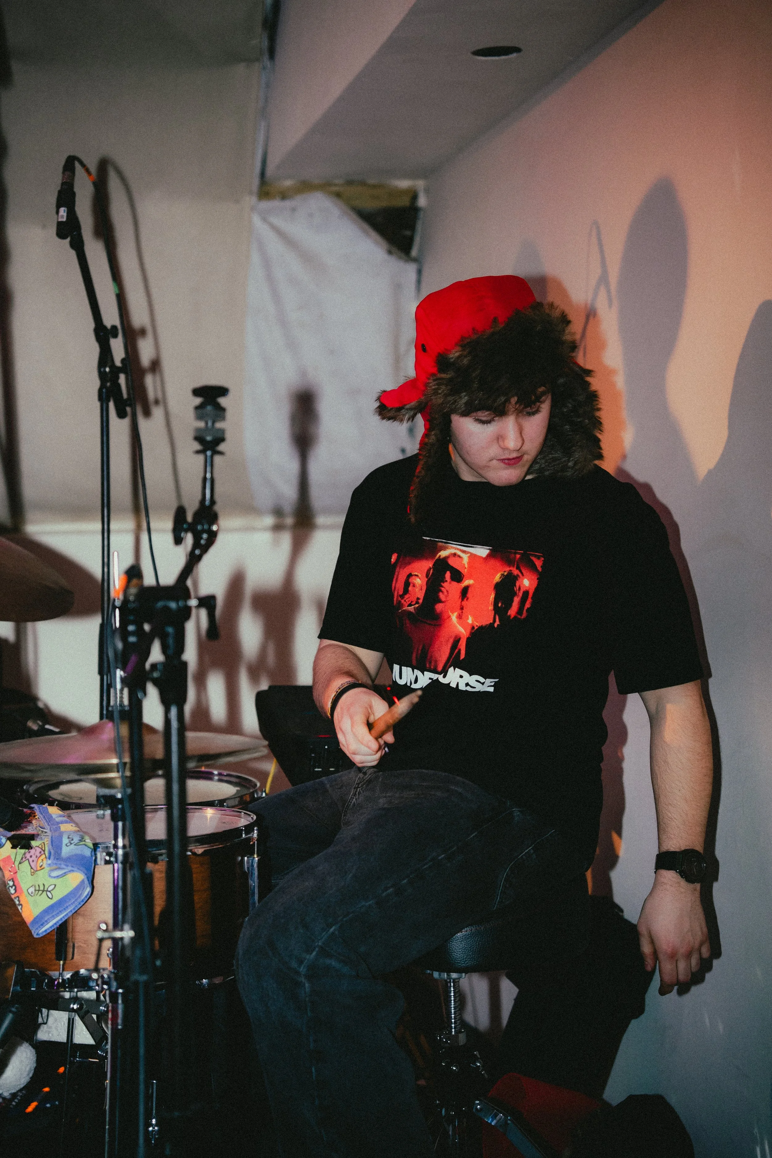 A young man wearing a red hat with fur, a black graphic t-shirt, and black jeans sitting on a drum stool holding a drumstick in a room with musical equipment.