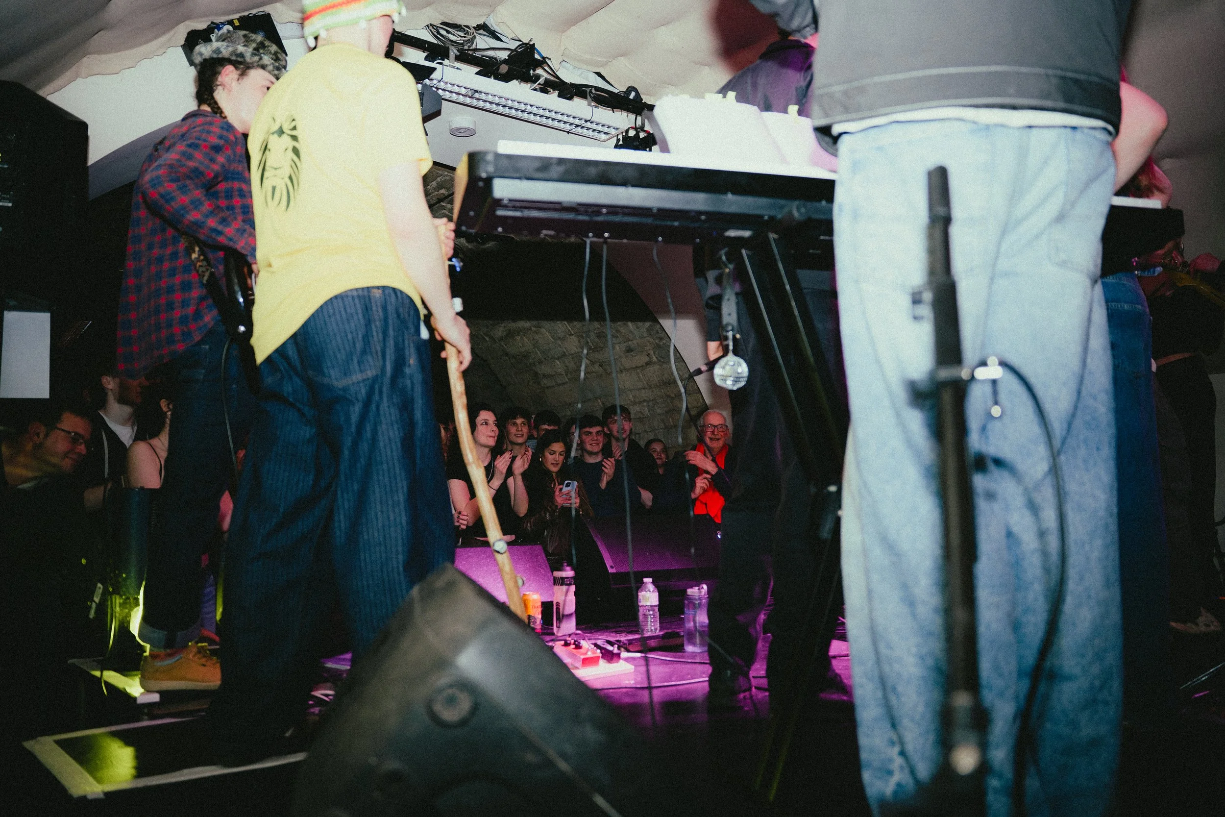 A live music or DJ performance is happening at a small indoor venue with a crowd of people watching and clapping.