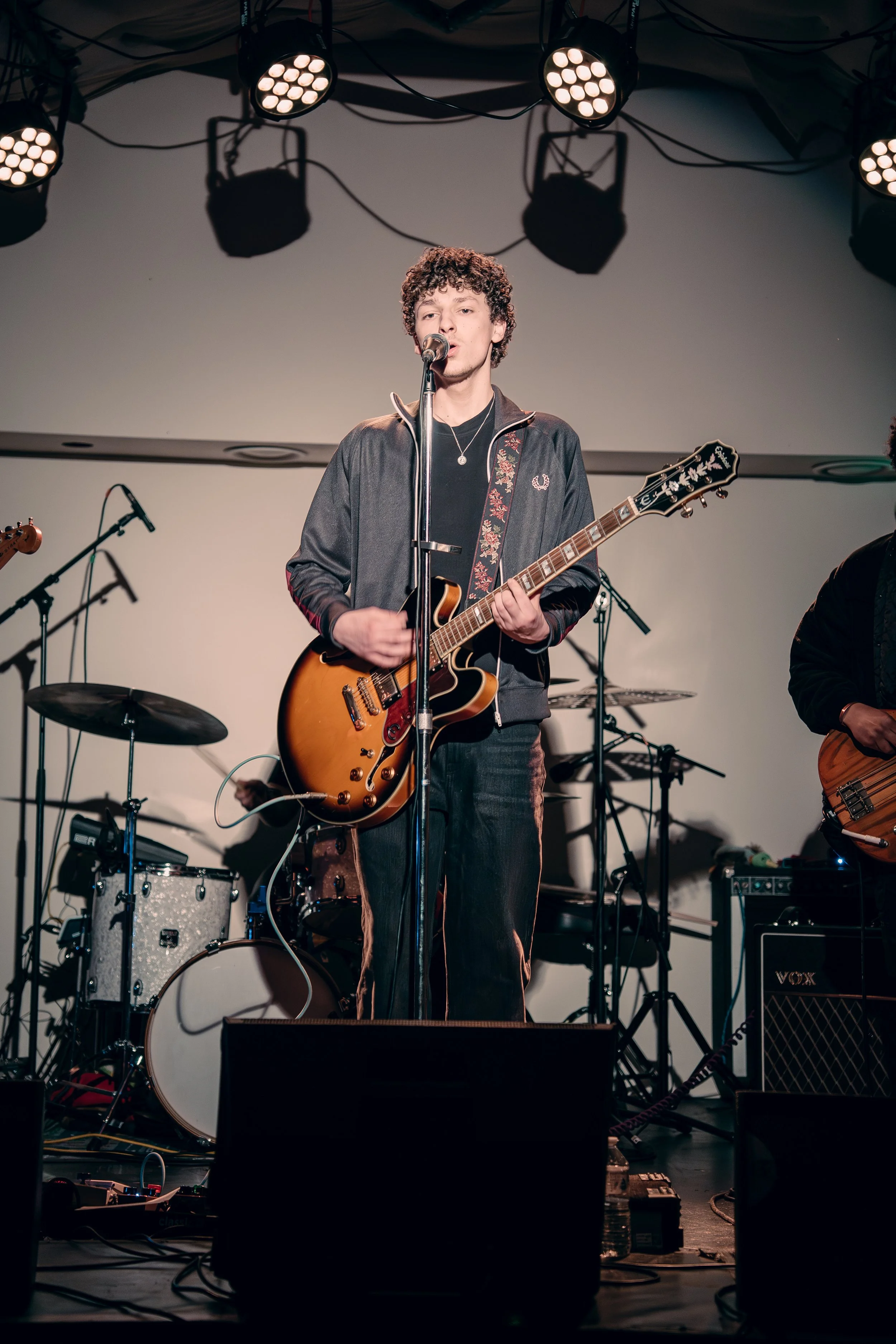 A young man with curly hair singing into a microphone while playing an electric guitar on stage during a live music performance.