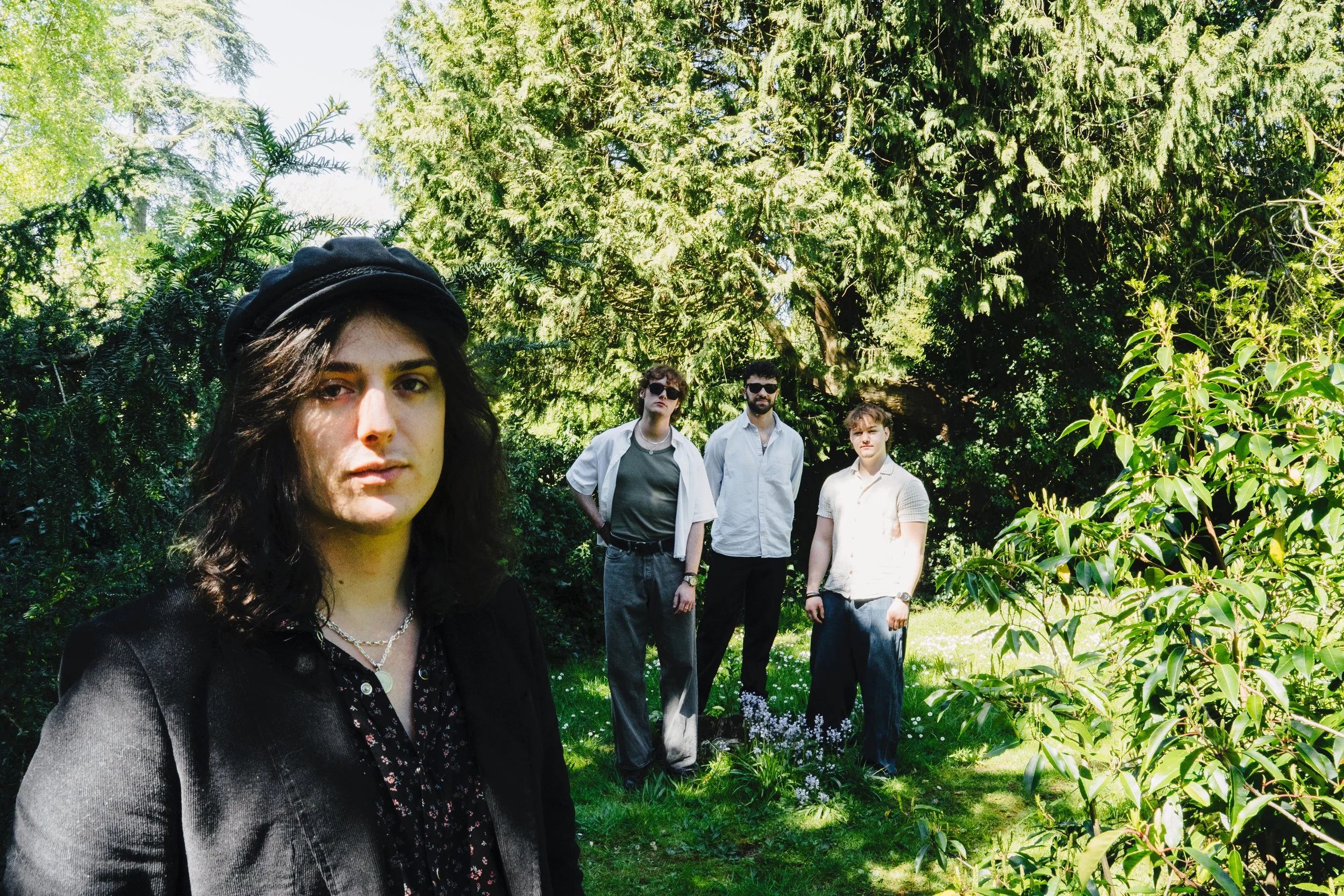 A group of five young adults standing outdoors in a lush green park with trees and bushes. One woman with dark hair and a black hat is in the foreground, with four men in the background.