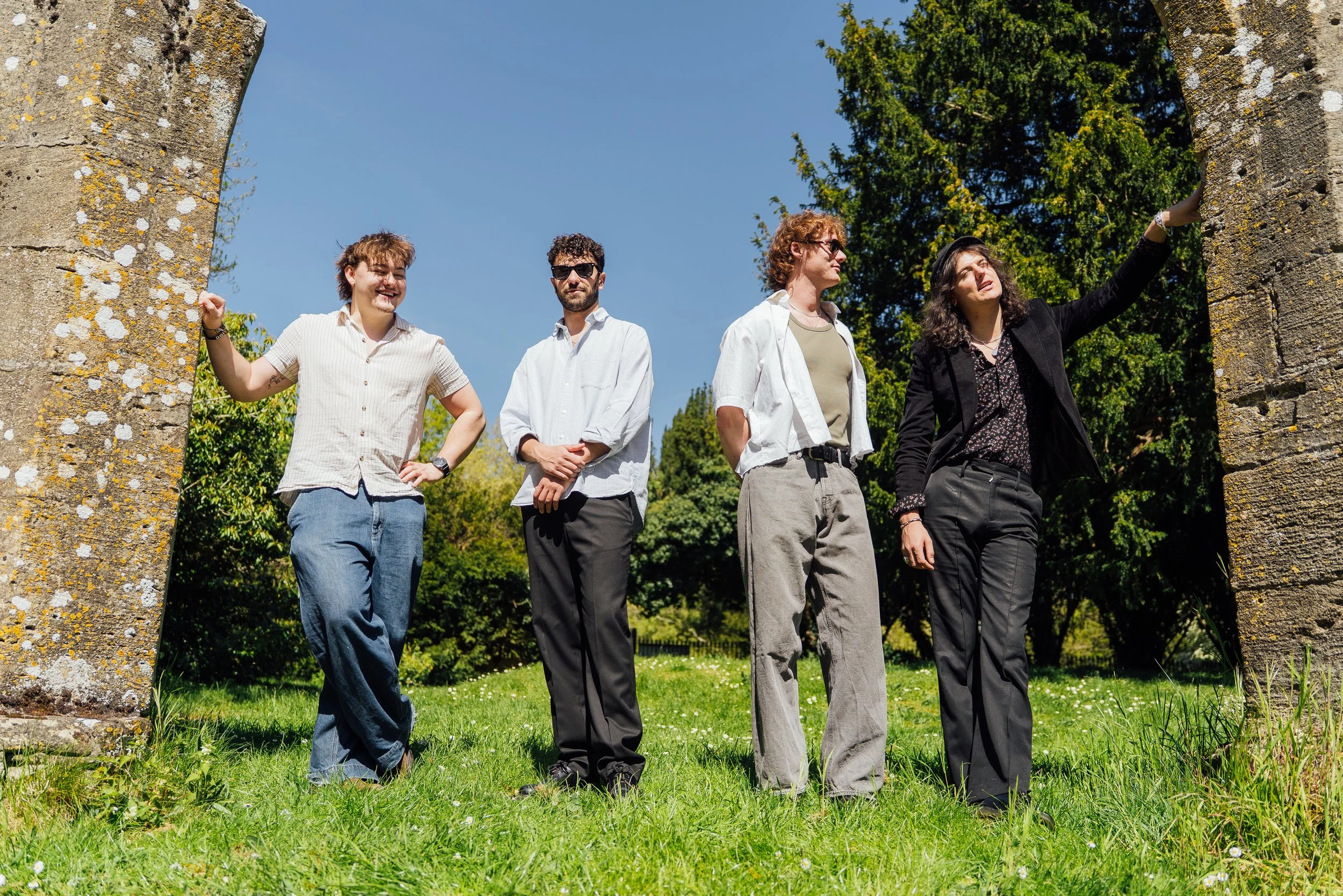 Four young adults standing outdoors between two stone columns, on a sunny day with green trees in the background.