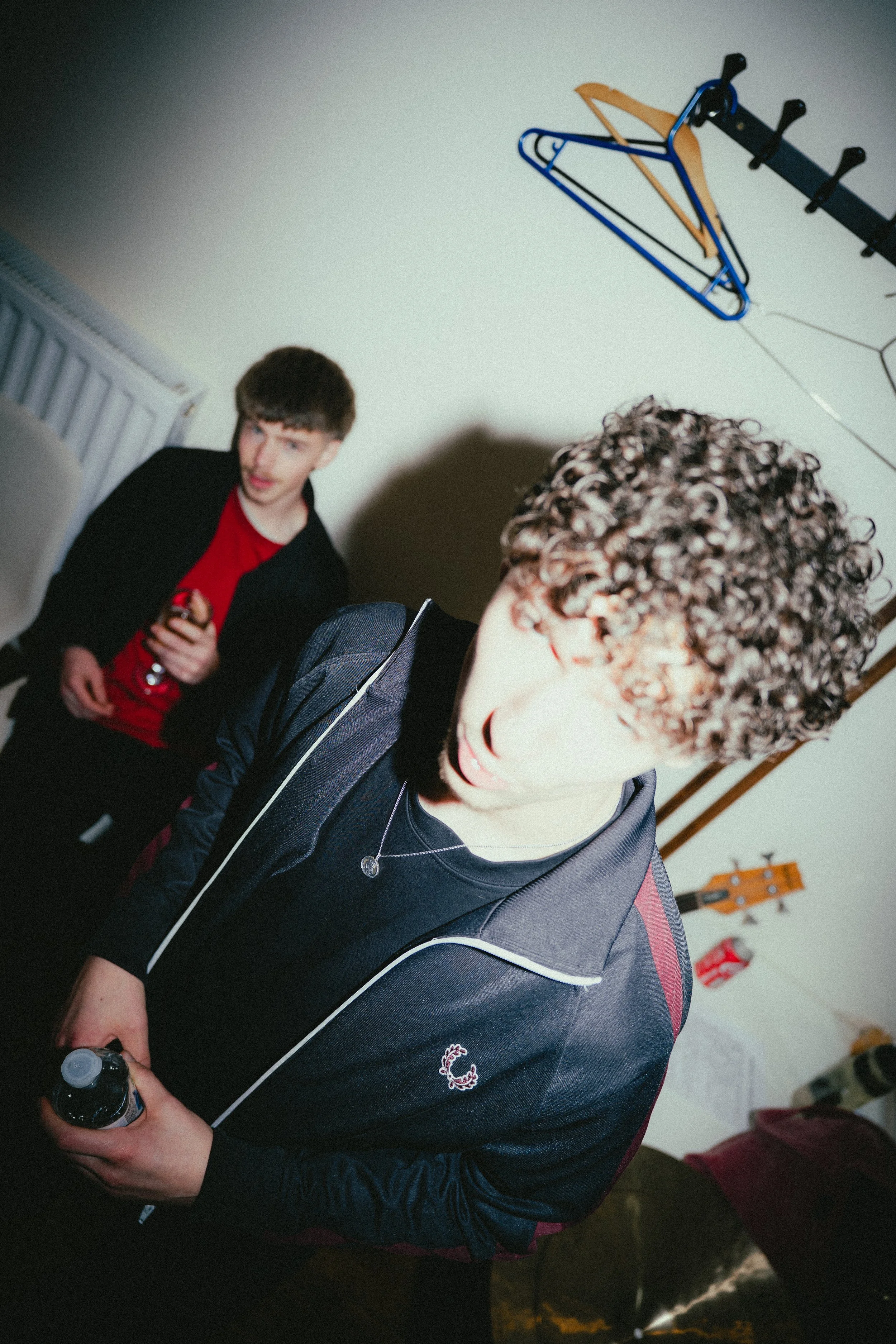 Two young men in a room, one with curly hair and holding a bottle, the other with straight hair and holding a drink, with clothes hangers and a guitar on the wall.