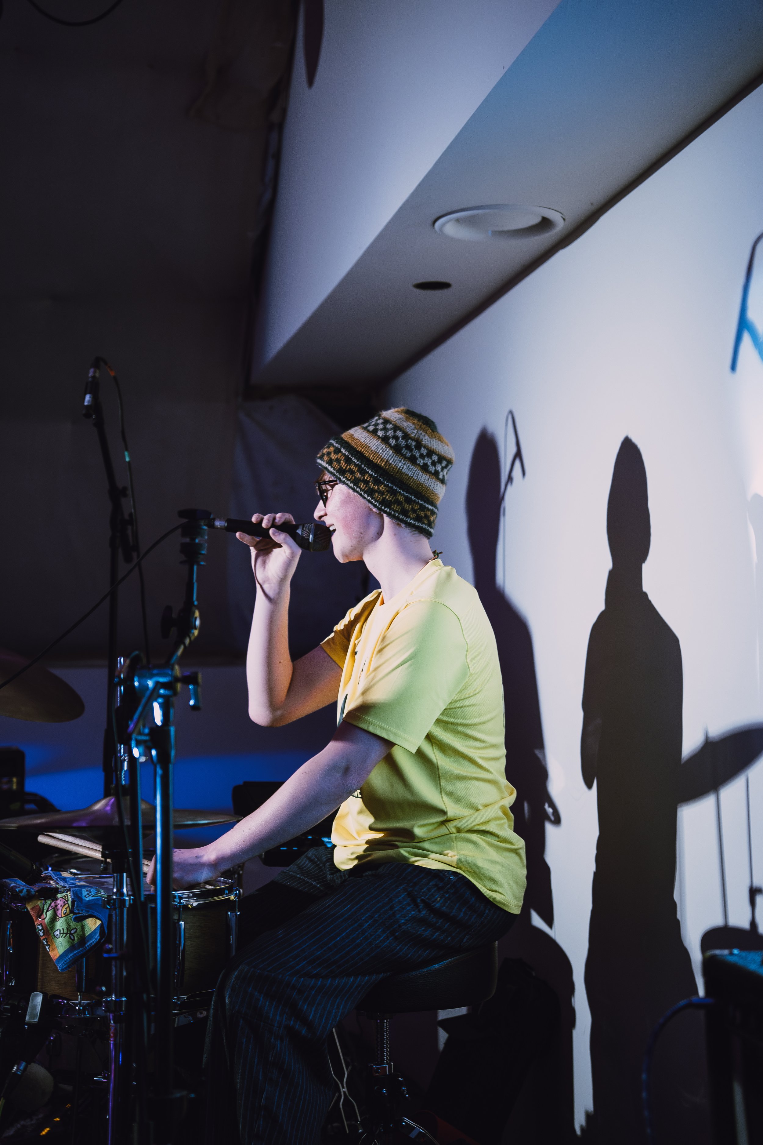 A woman wearing a yellow shirt, striped pants, and a knit cap with glasses is sitting on a stool, singing into a microphone, with a drum set nearby. Shadows of her and other objects are cast on a white wall behind her.