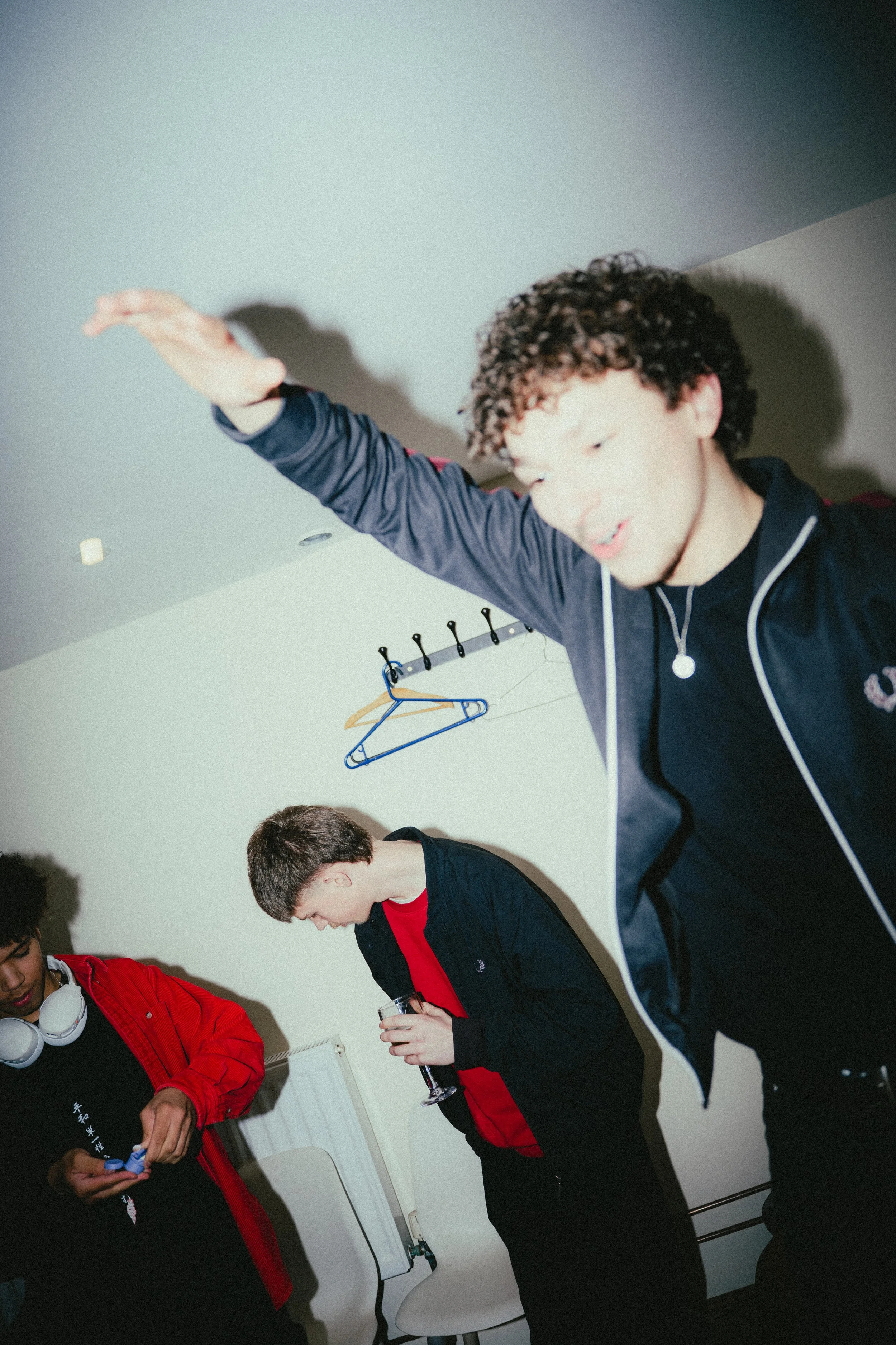 Three young men in casual clothing, two in black and one in red, are standing in a room with white walls and hooks on the ceiling. One is reaching upward, and the others are looking at their phones, holding drinks.