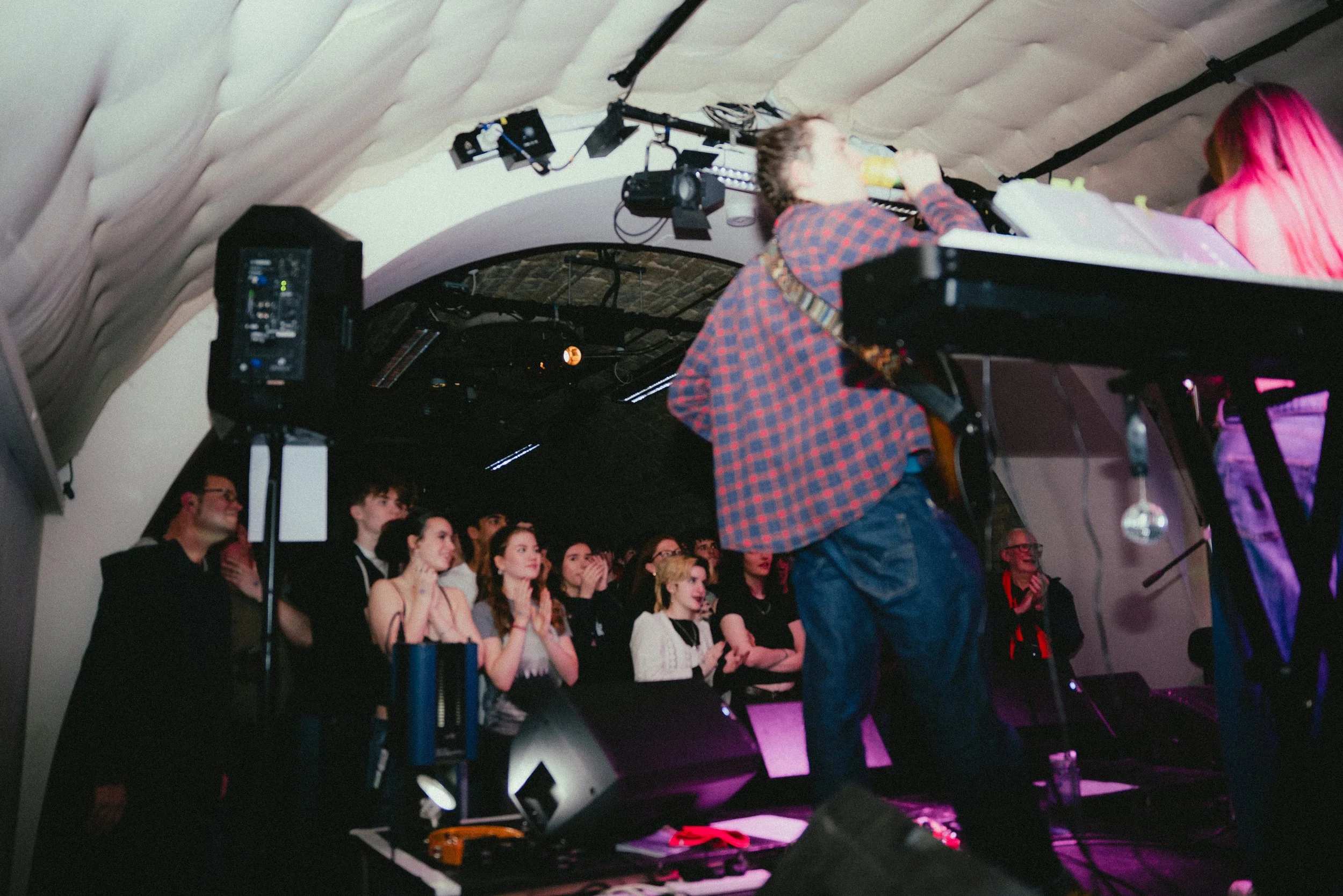 A performer on stage with a guitar singing into a microphone while an animated audience watches, some with expressions of surprise and admiration, in a dark, indoor venue.