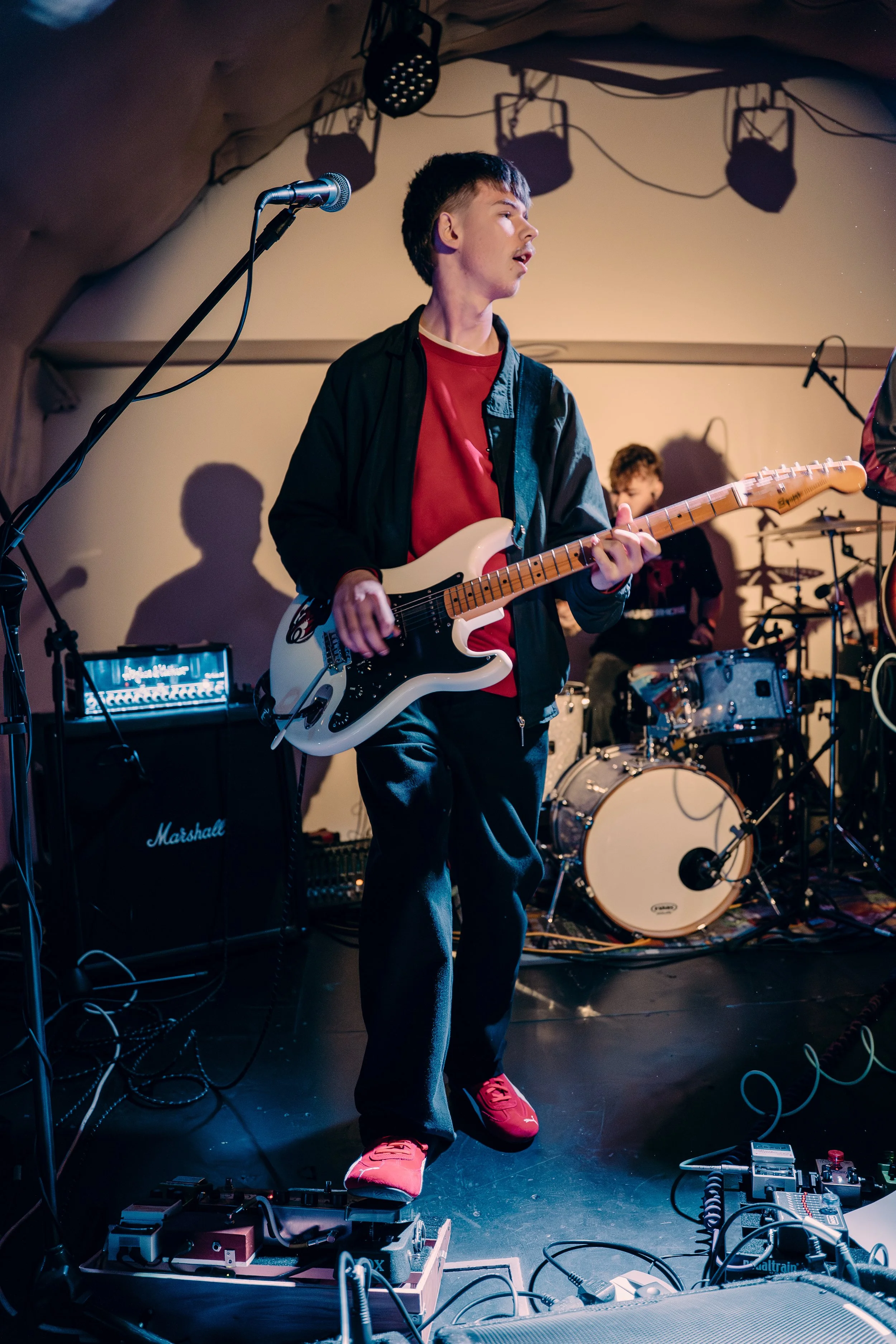 Young male musician playing an electric guitar on stage with a drum set behind him, performing at a live concert.