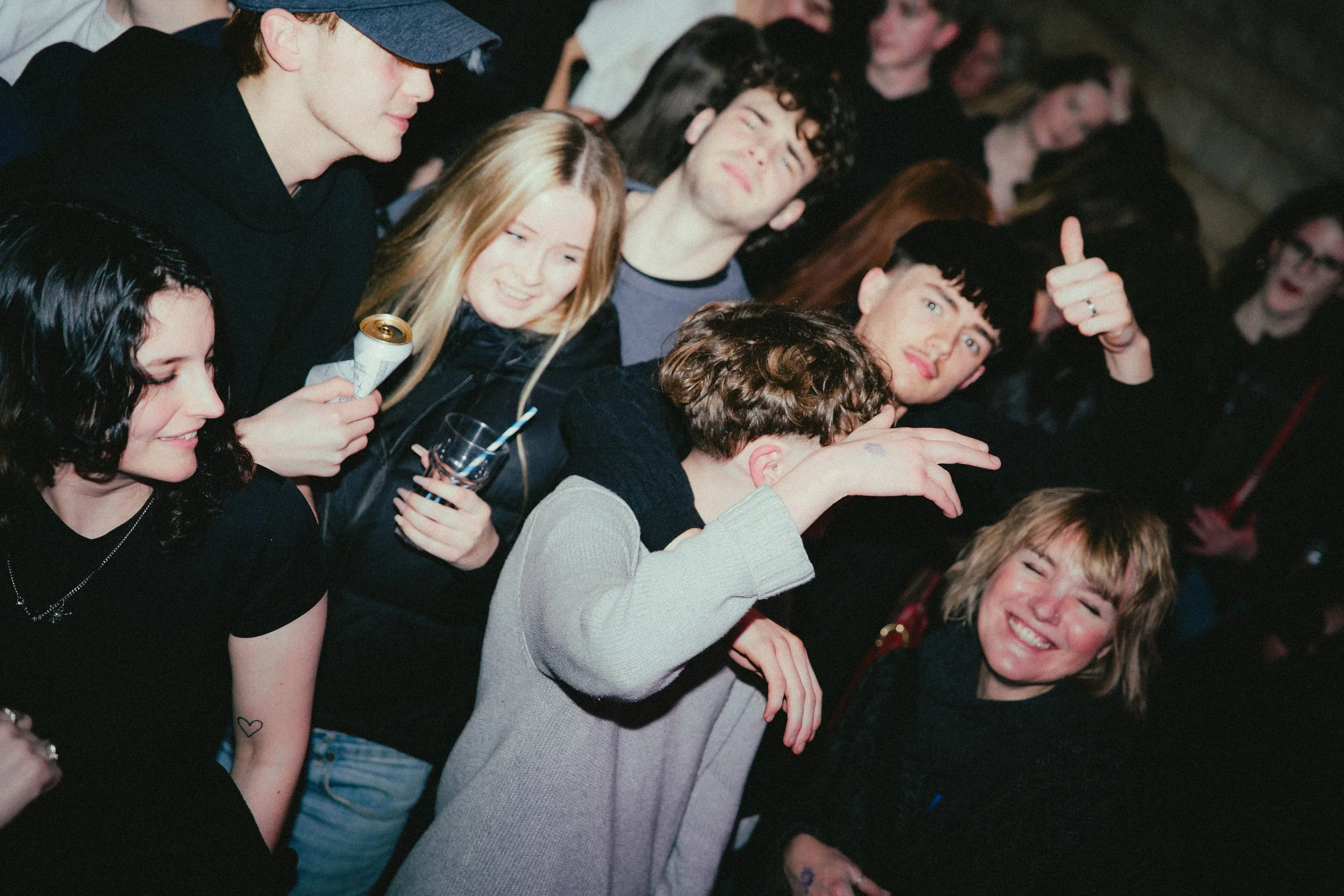 Group of young people socializing indoors at night, smiling, laughing, some holding drinks, casual attire.