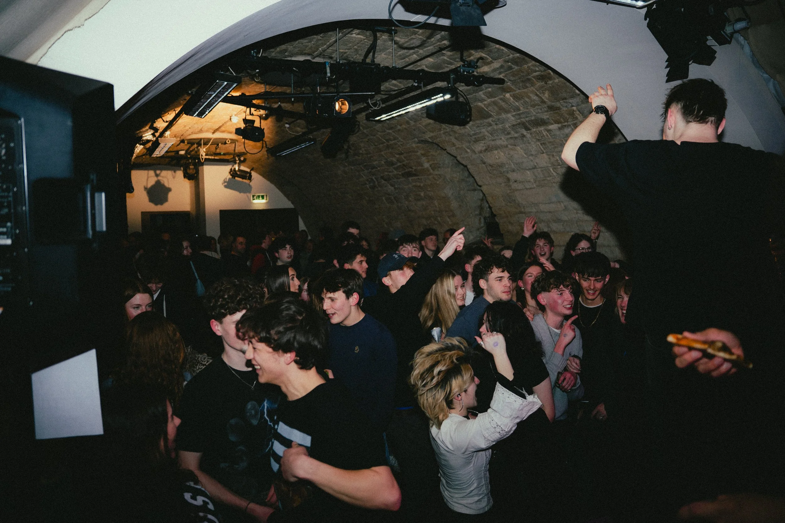 A crowded indoor music venue with people dancing and enjoying a live performance. The venue has a stone arched ceiling and stage lighting equipment hanging from above. A performer is on stage with their back to the camera, engaging with the audience.