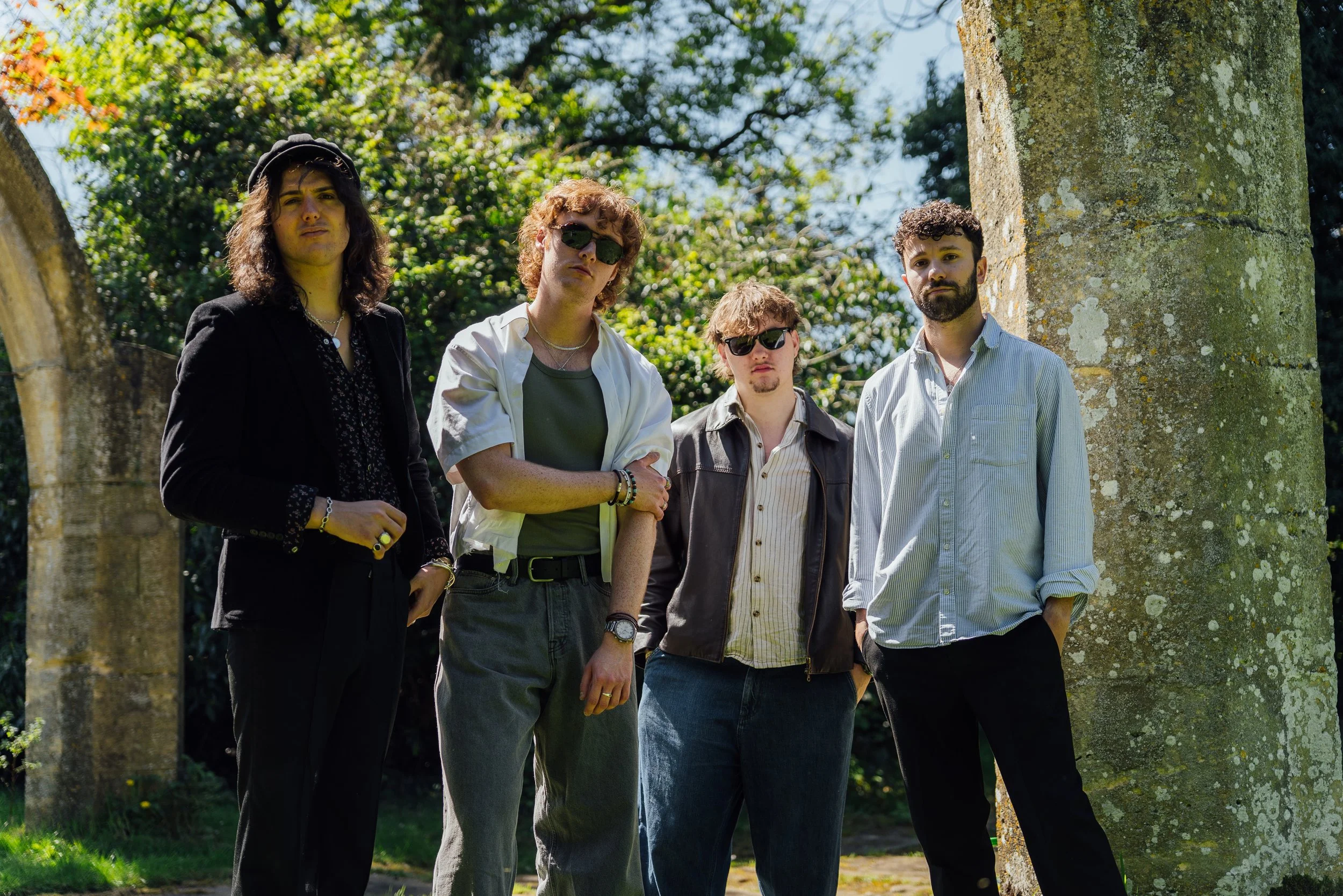 Four young men standing outdoors near a stone pillar, with trees and greenery in the background. They are dressed casually, with some wearing sunglasses and jewelry, looking directly at the camera.