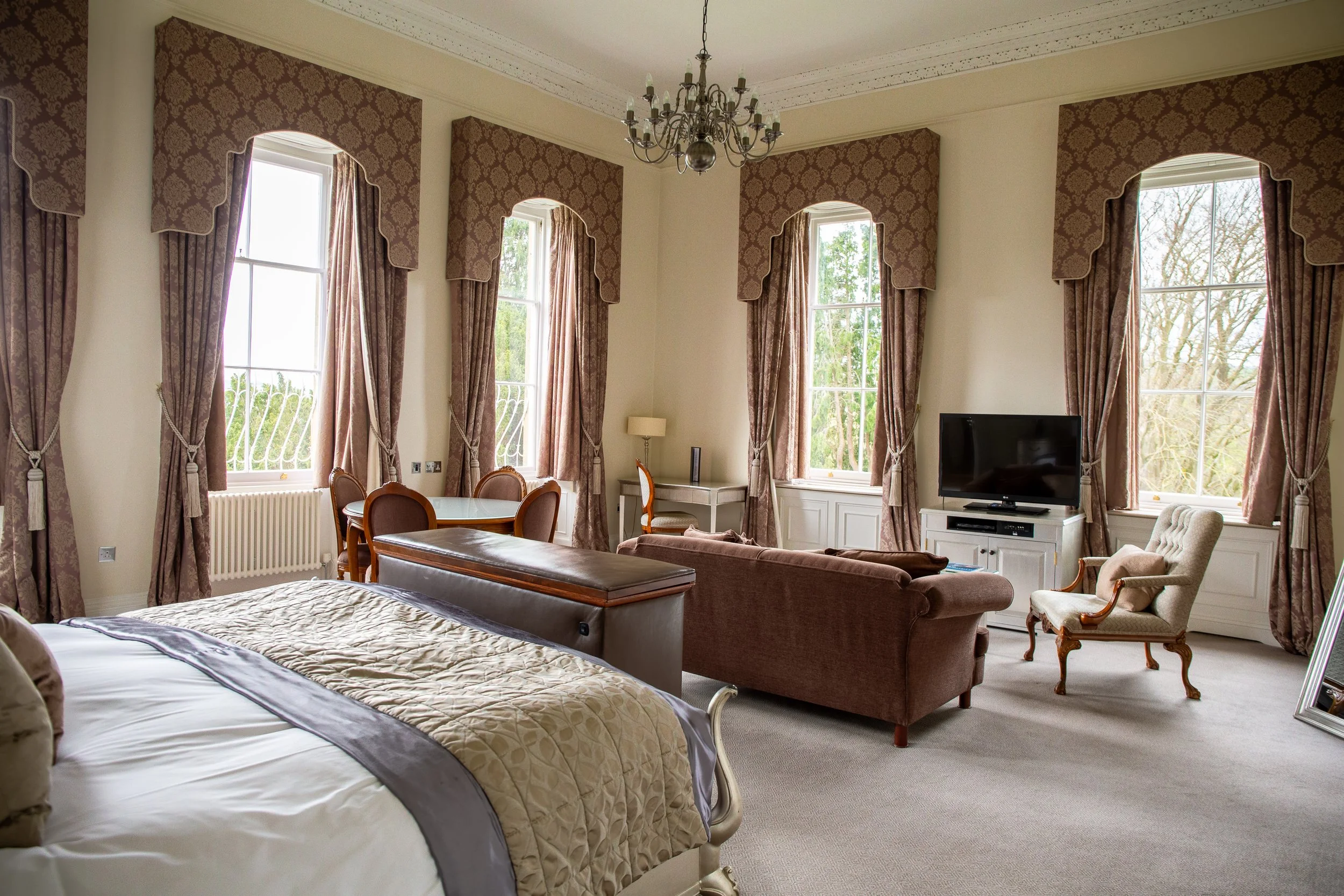 Elegant bedroom with large windows, draped curtains, a bed, a sitting area with a sofa and armchair, a television, and a chandelier.