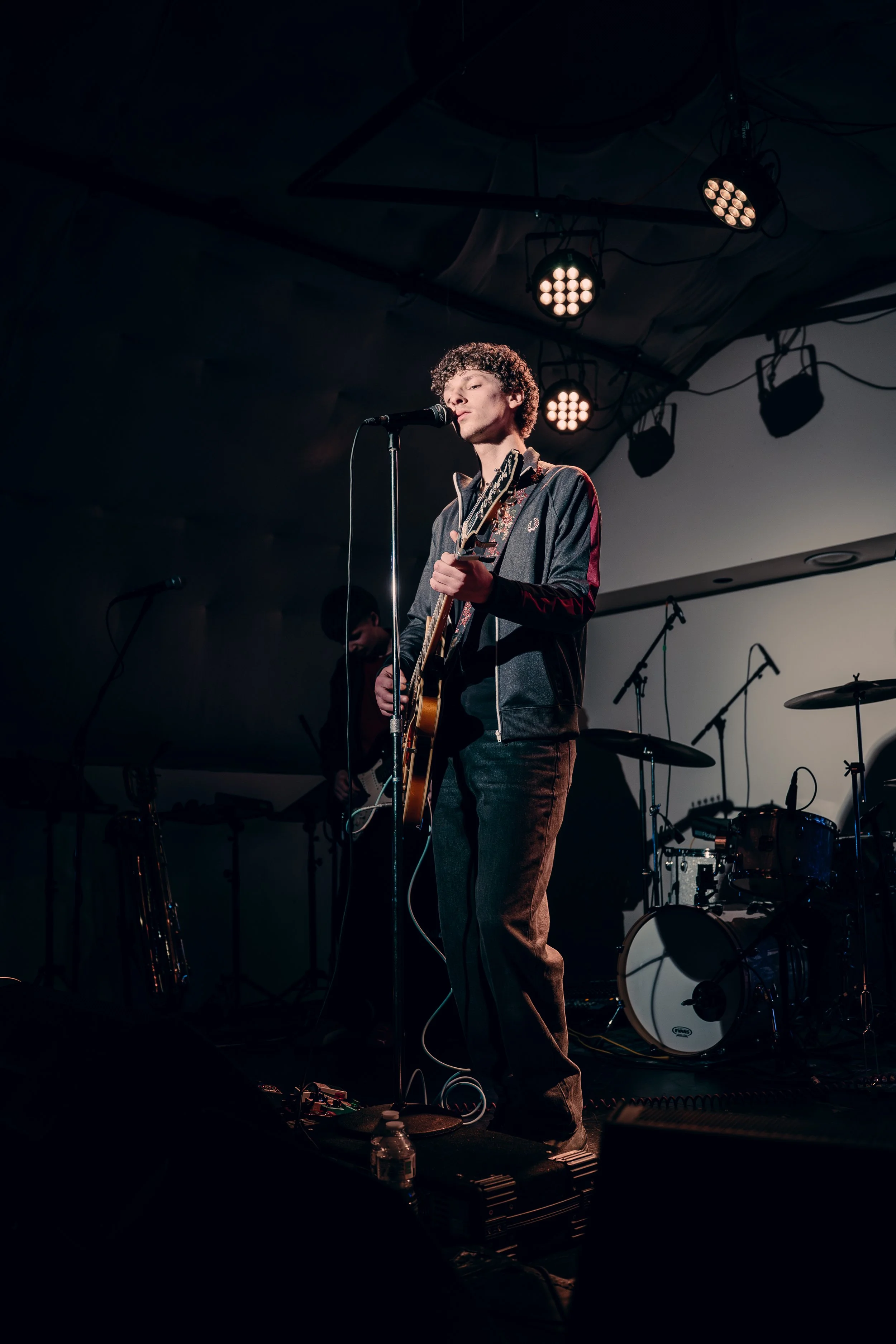 A male musician with curly hair wearing a dark jacket playing an acoustic guitar on stage, with a drum set and other musical equipment in the background, under stage lighting.