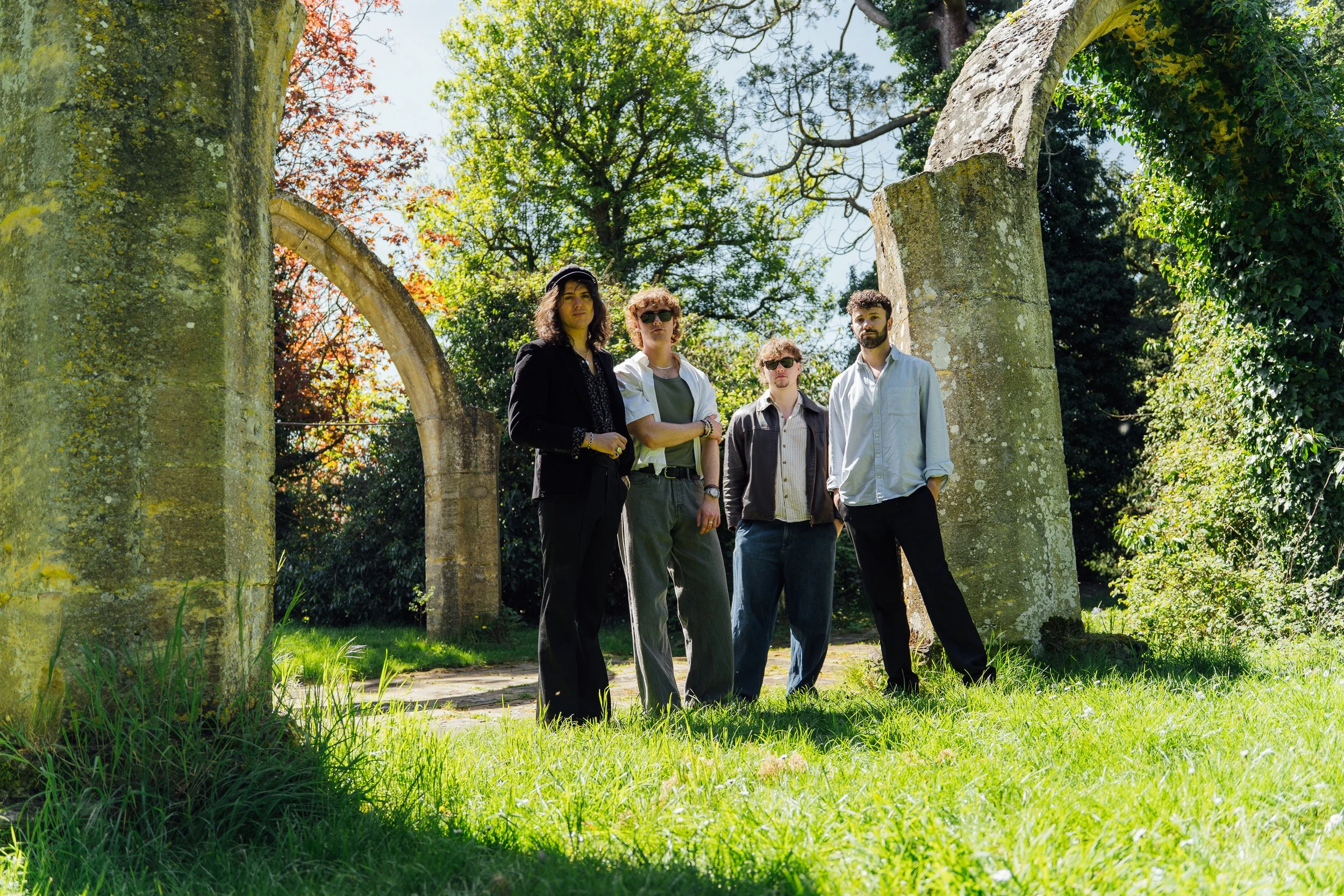 Four young adults standing together outdoors among ancient stone ruins and lush green trees, with bright sunlight filtering through the leaves.