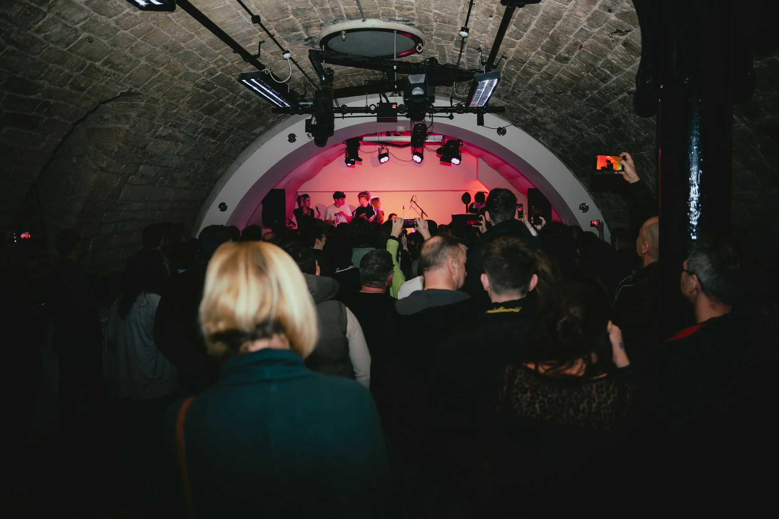 People watching a live musical performance in a small, dimly lit venue with a curved brick ceiling and stage lighting.