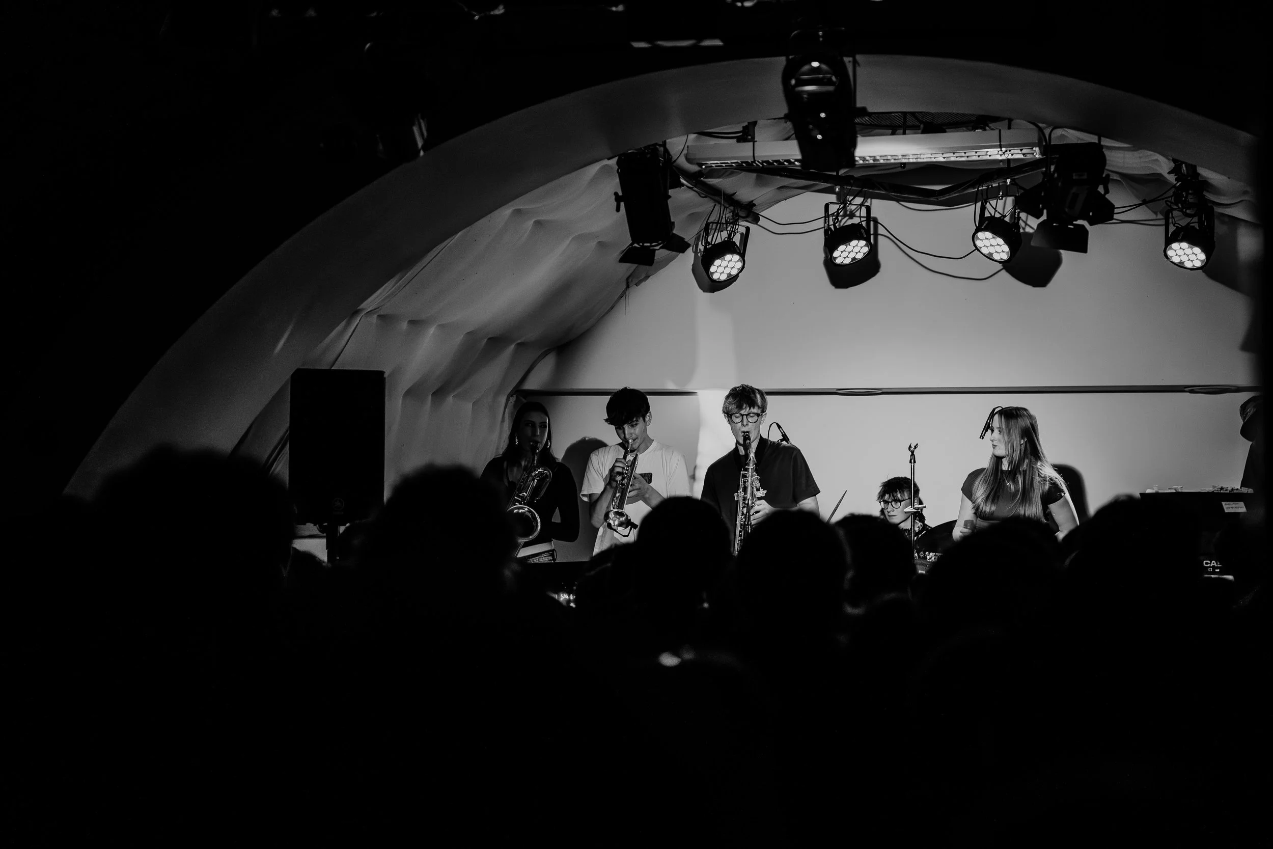 A black and white photo of a live band performing on stage with an arched ceiling. The band includes musicians playing saxophones and a person on drums. Audience silhouettes are visible in the foreground.
