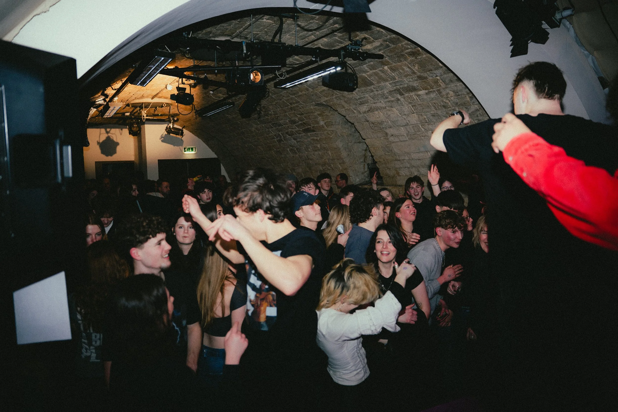 A crowd of young people dancing and enjoying themselves at a concert or nightclub, with a brick arched ceiling overhead.