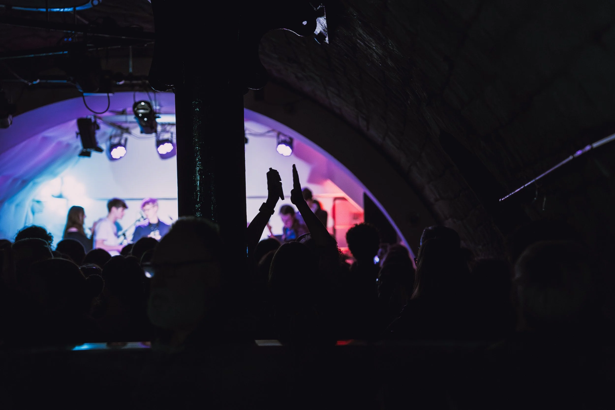 Silhouettes of people in a darkened concert venue with a stage in the background illuminated by colorful lights, and two people in the crowd raising their hands.