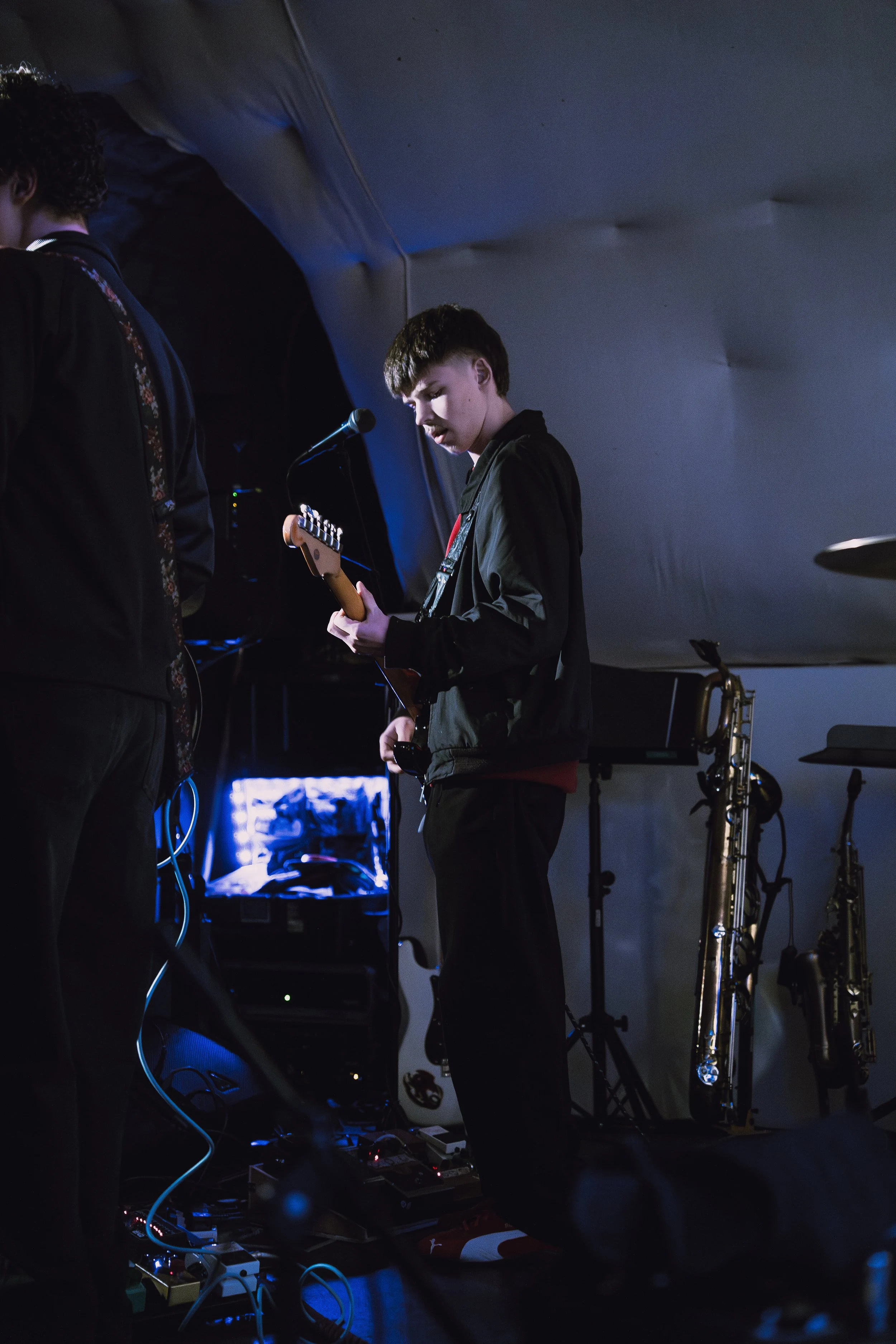 Young man with short hair playing an electric guitar on a dimly lit stage, surrounded by musical equipment and saxophones in the background.