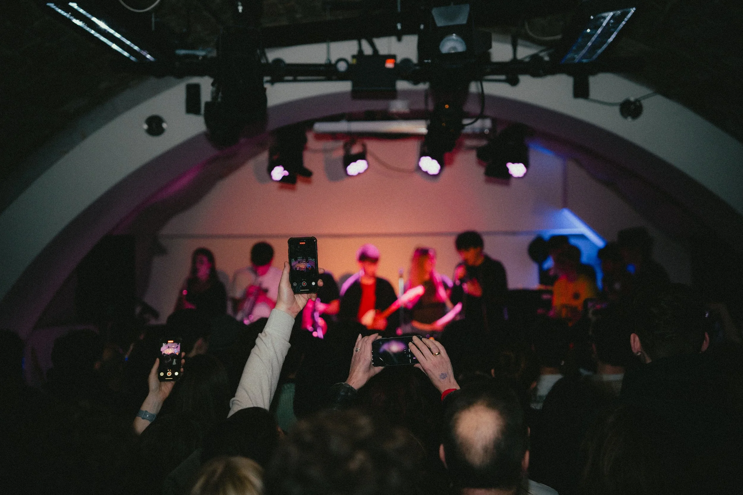 People taking photos and videos of a stage performance at a concert or event, with performers on stage under colorful lighting.