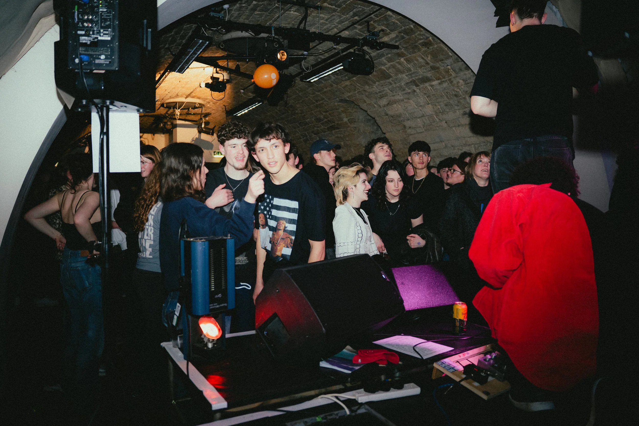 Crowd of young people at a music event or concert in a dimly lit venue with arched brick ceiling, with sound and stage equipment visible.