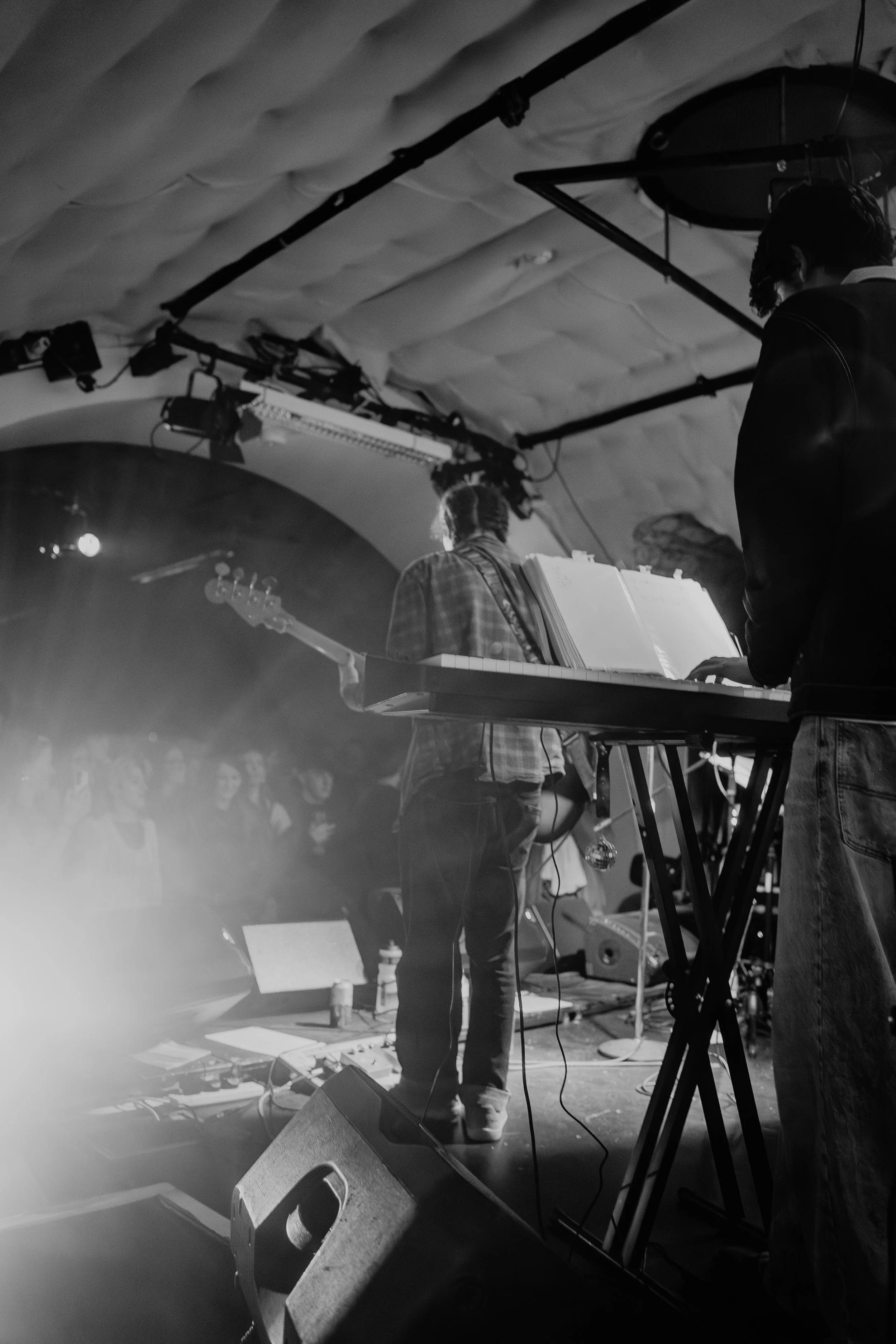 Black and white photo of musicians performing on stage, with a keyboard and a guitar visible, audience in the background.