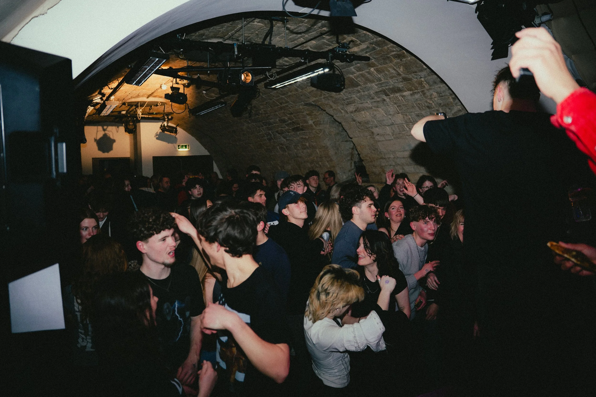 A crowd of young people enjoying a concert or DJ performance in an underground venue with an arched brick ceiling. Some people are dancing, talking, or cheering.