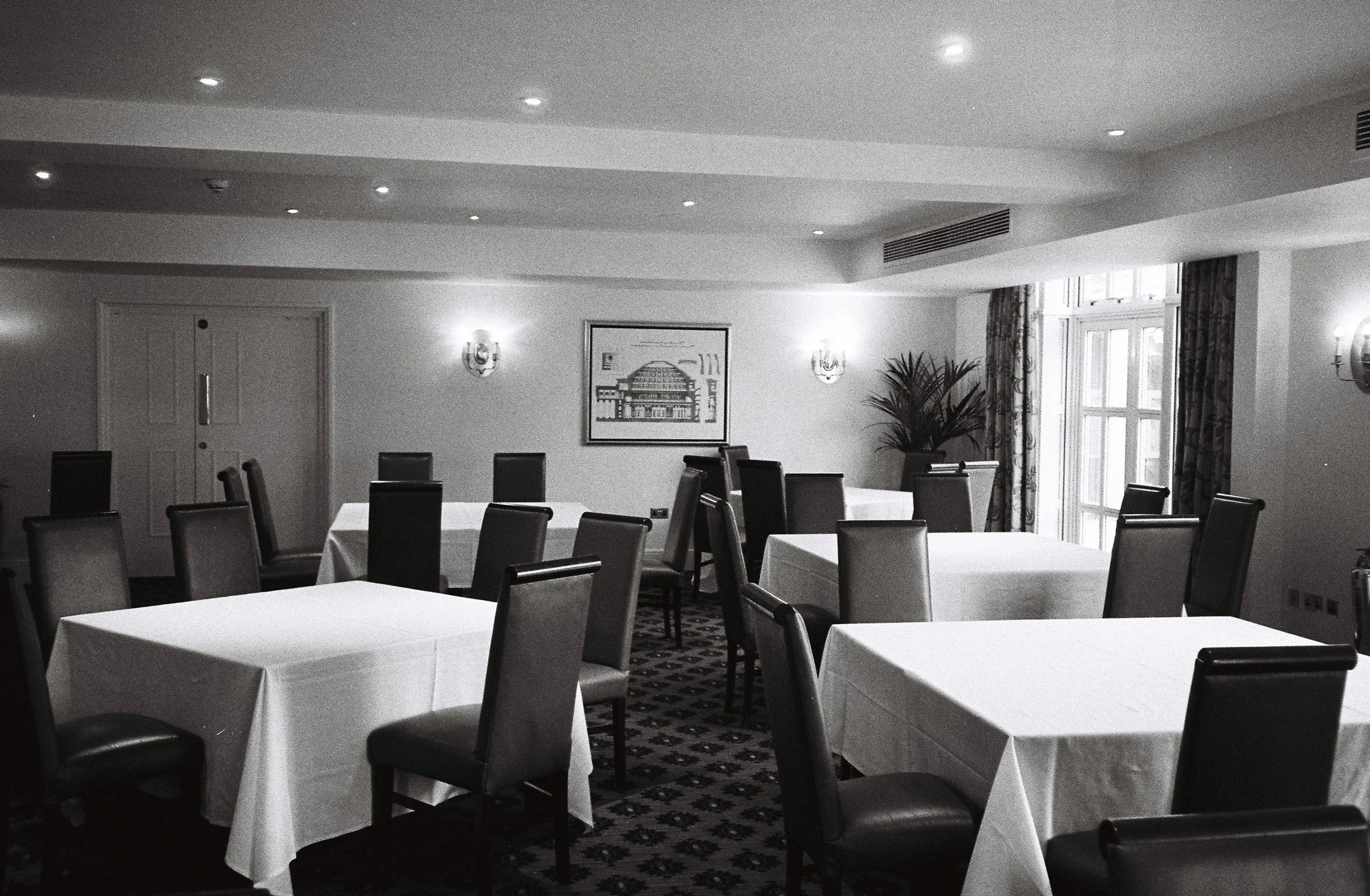 A conference room with several tables covered in white tablecloths and surrounded by dark chairs, with a large window, potted plant, and framed picture on the wall.