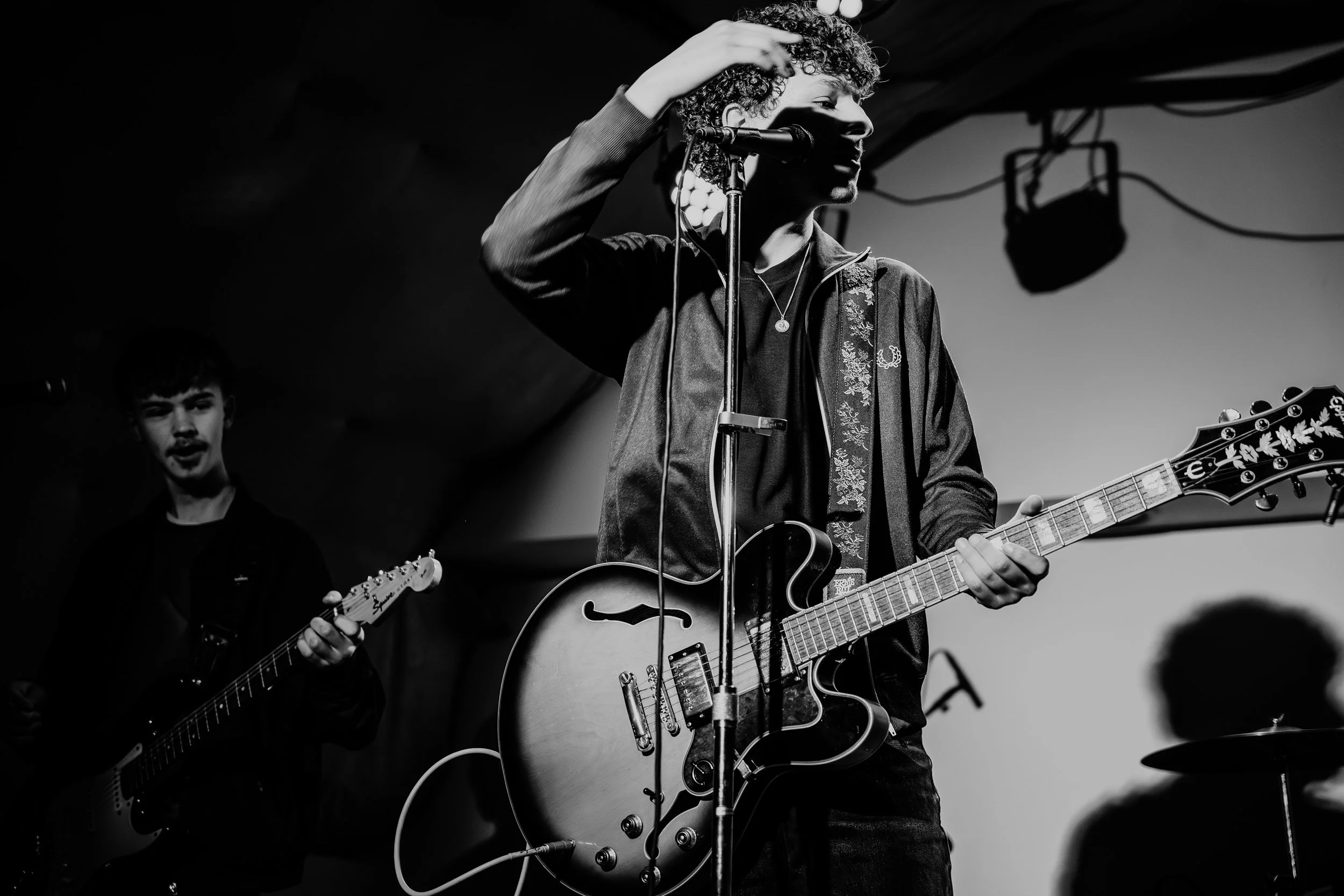 Black and white photo of a musician playing an electric guitar and singing into a microphone on stage, with another guitarist in the background.