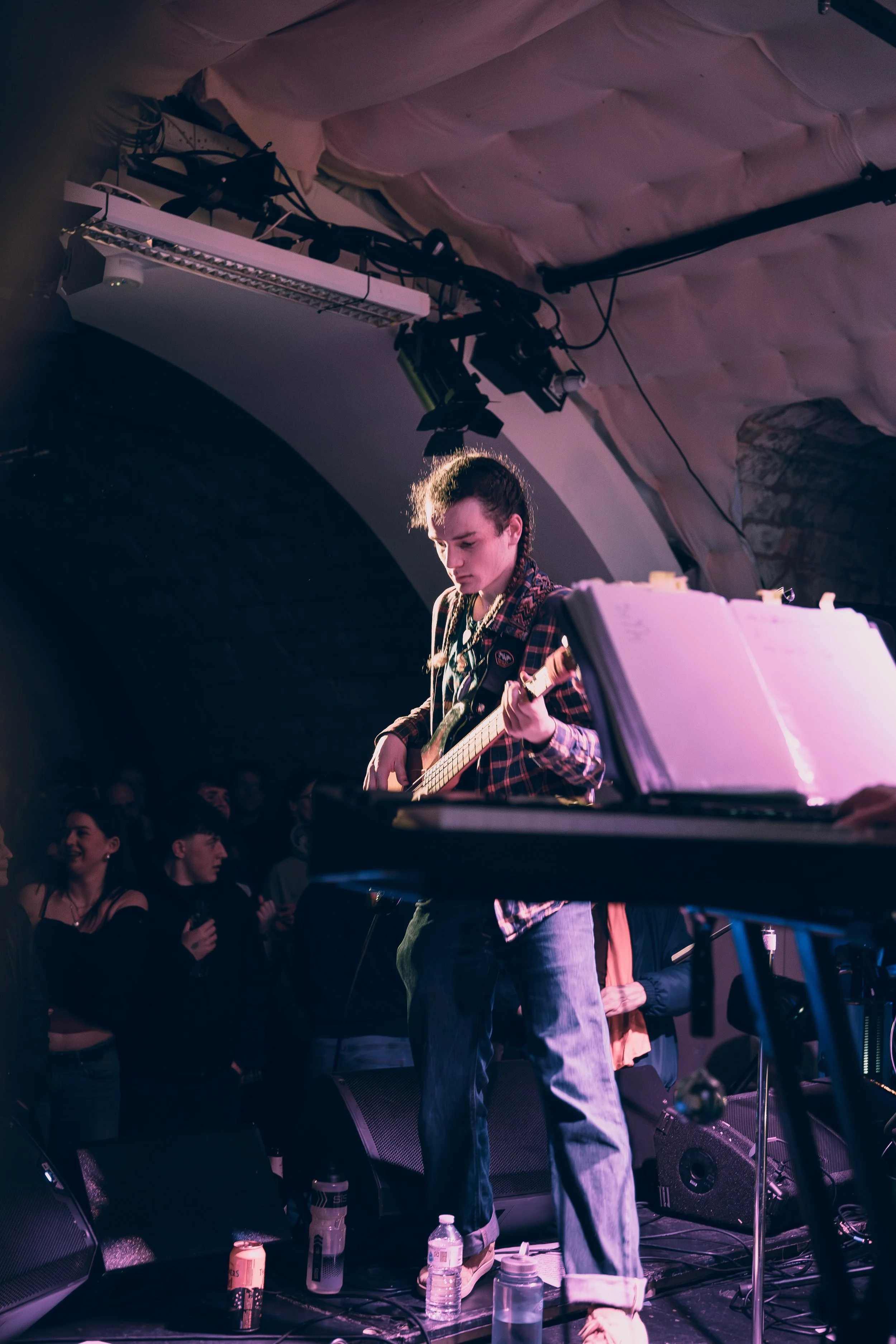 A young musician playing an electric guitar on stage at a concert venue with an audience in the background.