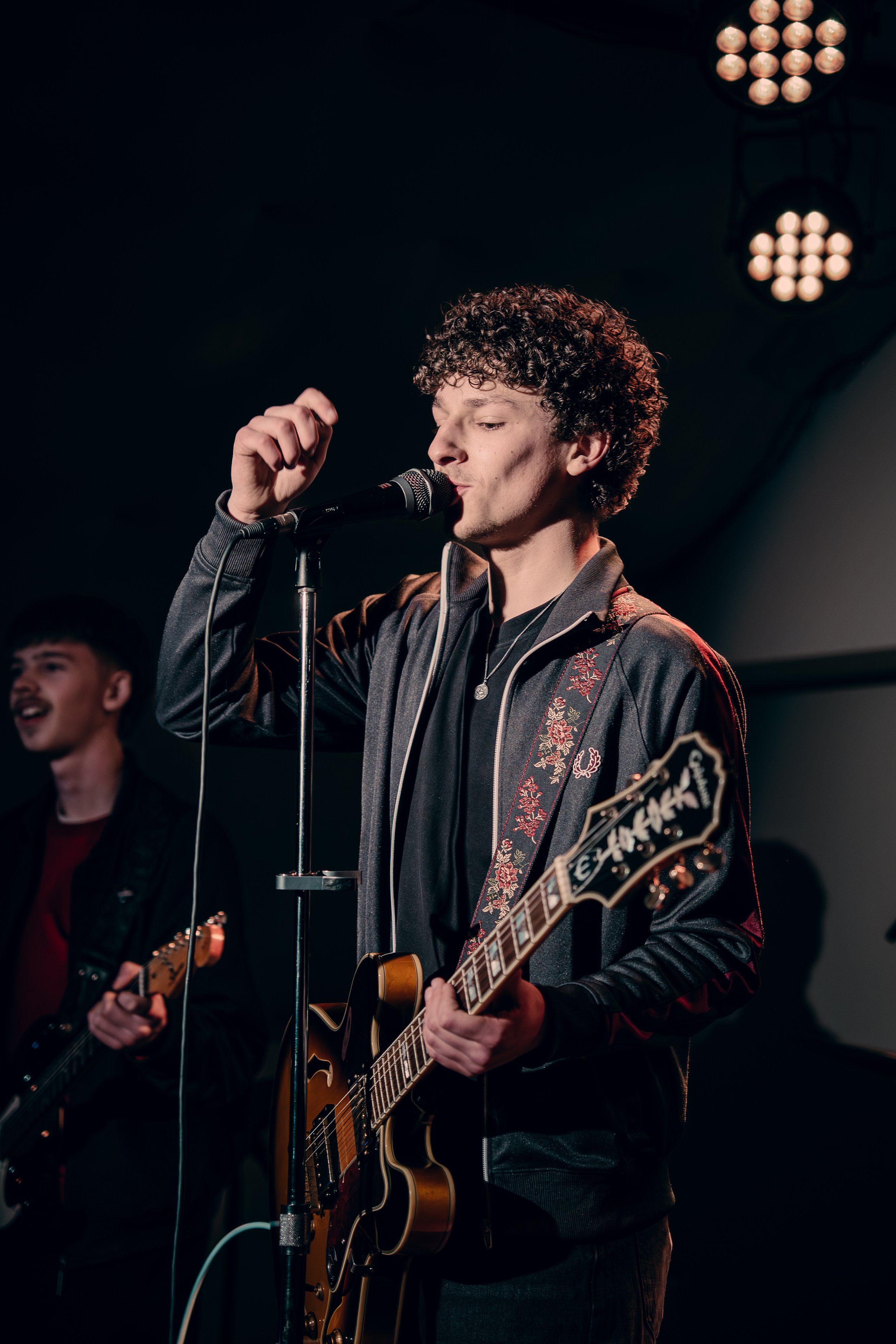 Young man with curly hair singing into a microphone while playing an electric guitar during a live performance.