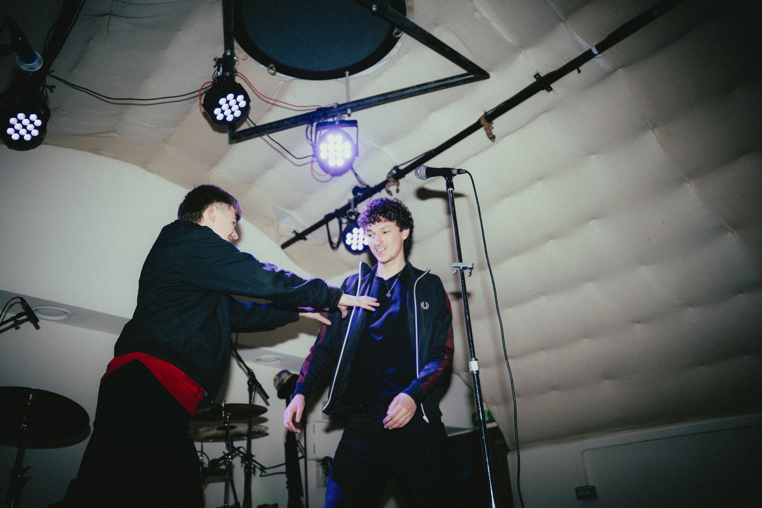 Two young men on stage, one smiling and the other reaching toward him. Microphones and stage lights are visible above them.
