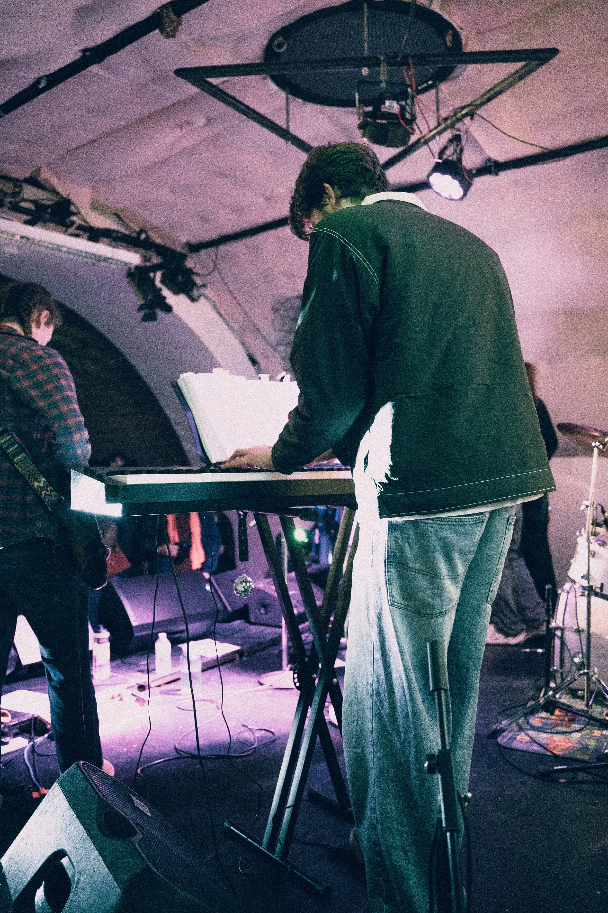 Musicians performing on stage, with one focusing on a keyboard, in a concert venue with stage lighting and equipment.