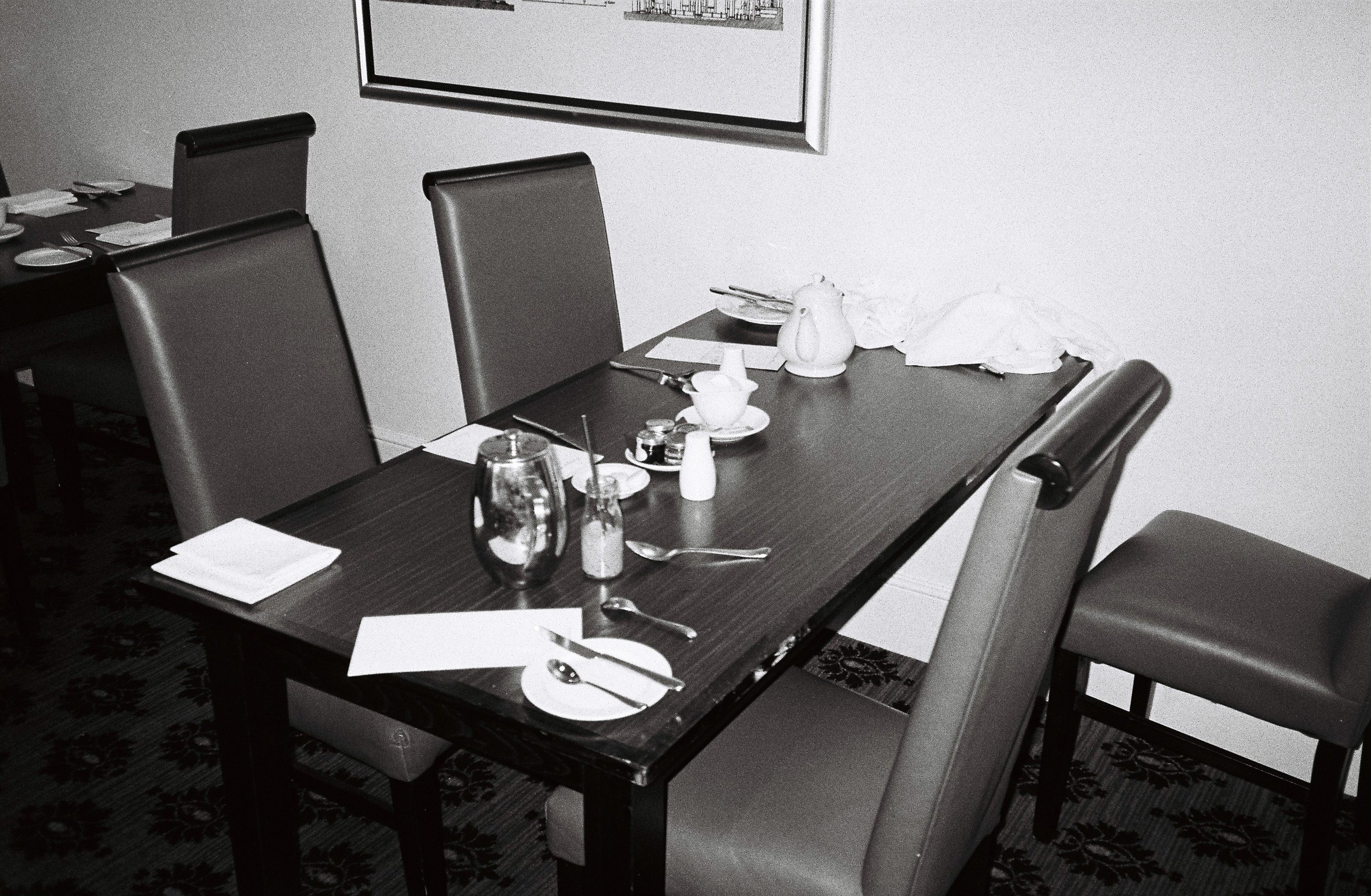 A formal dining table set for a meal, with plates, silverware, napkins, a glass, salt and pepper shakers, and a pitcher, in a room with patterned carpet and framed artwork on the wall.