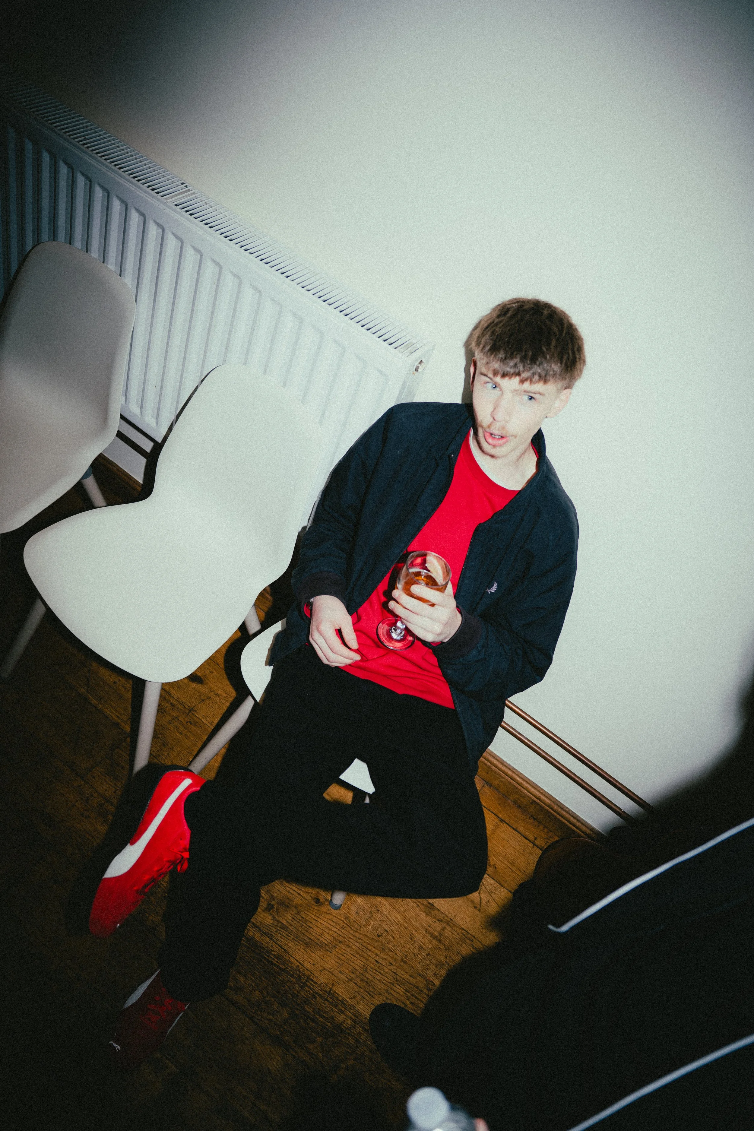 A young man sitting on a white chair, holding a glass of drink, dressed in red and black with red sneakers, making a surprised expression. There are additional chairs and a radiator behind him.
