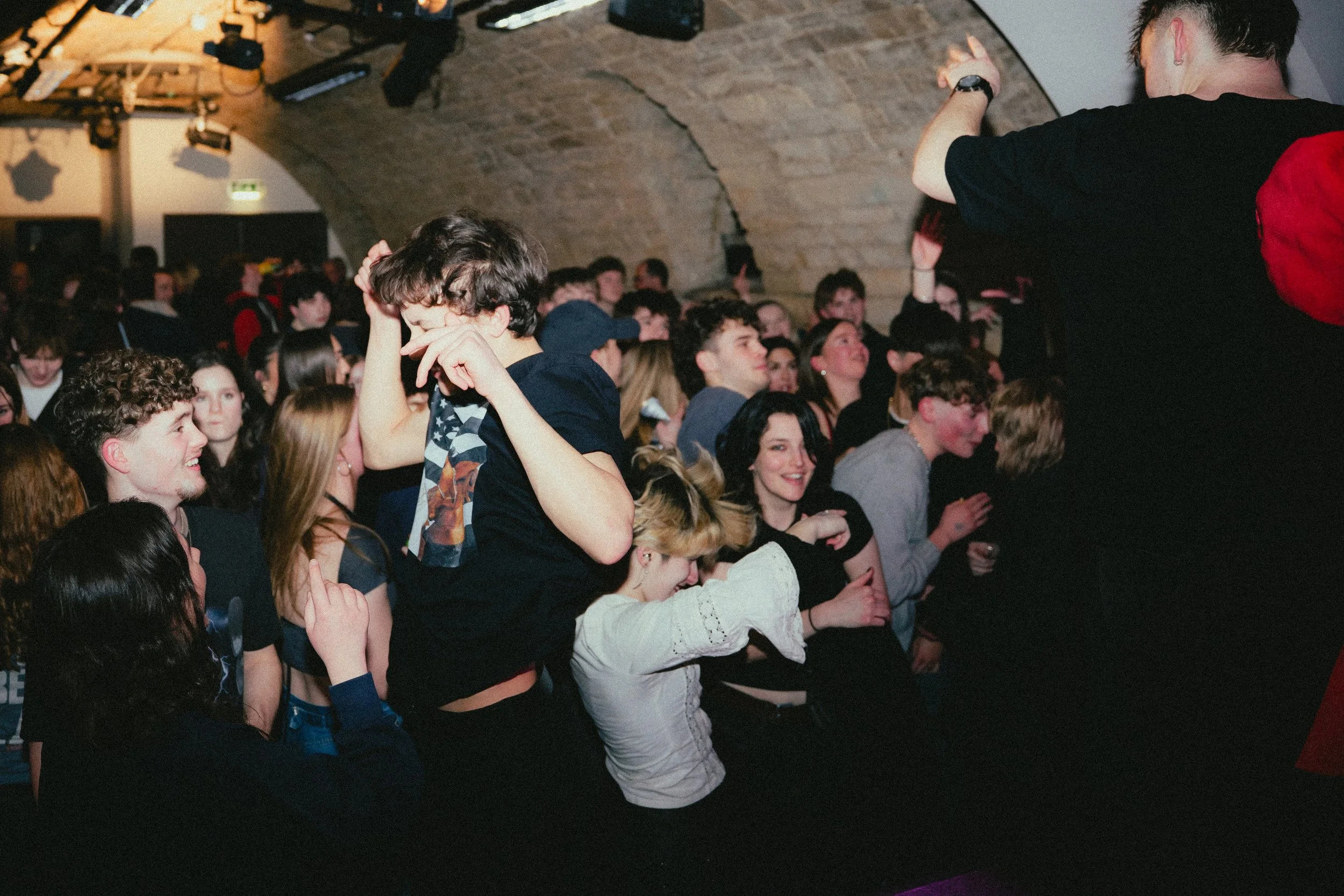 Crowd of young people at a concert or event, smiling and interacting, in a dimly-lit venue with brick walls.