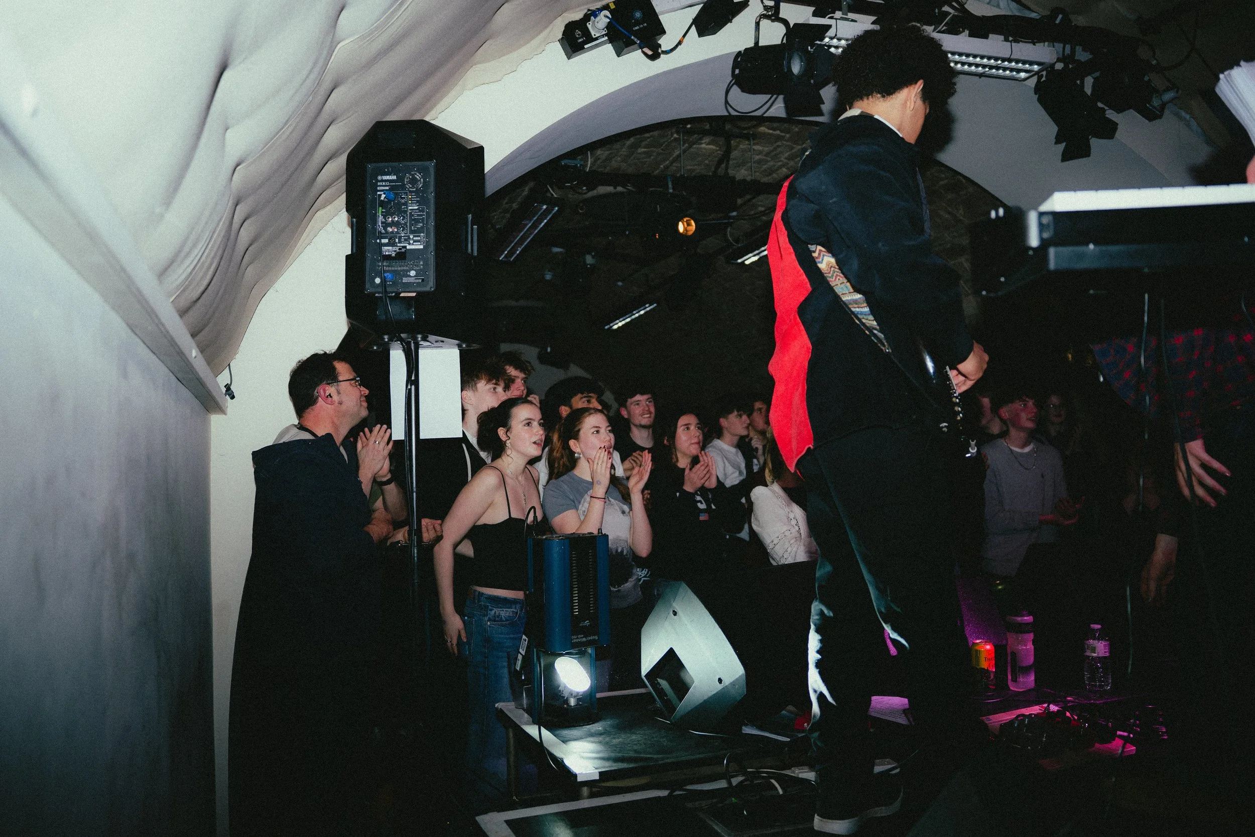 Audience of young people watching a performer DJ at a nightclub or concert. The crowd appears engaged, some clapping and cheering, under dim lighting.