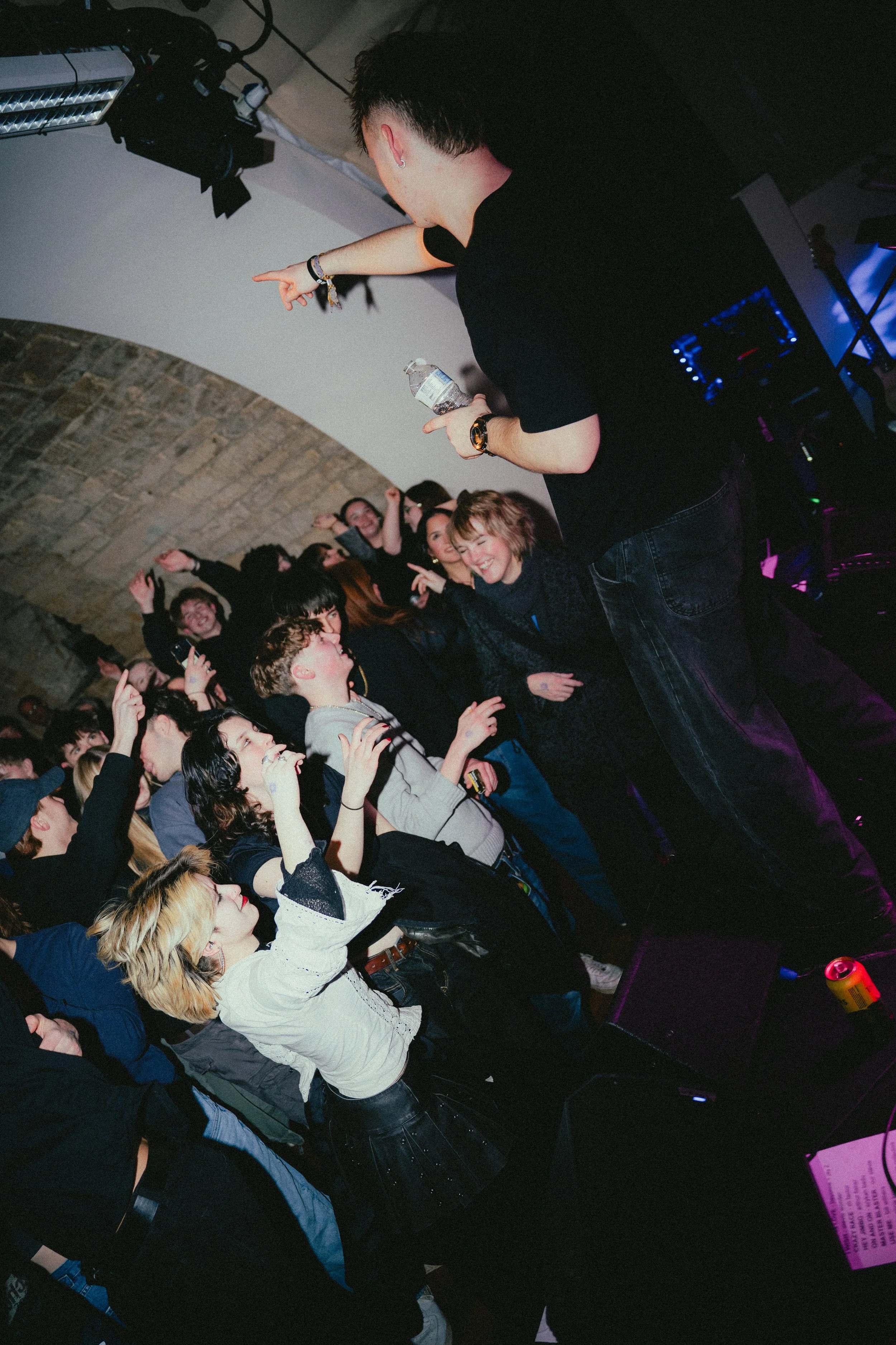 A DJ or performer on a stage interacting with a lively crowd in a nightclub or party setting, with people dancing and smiling.